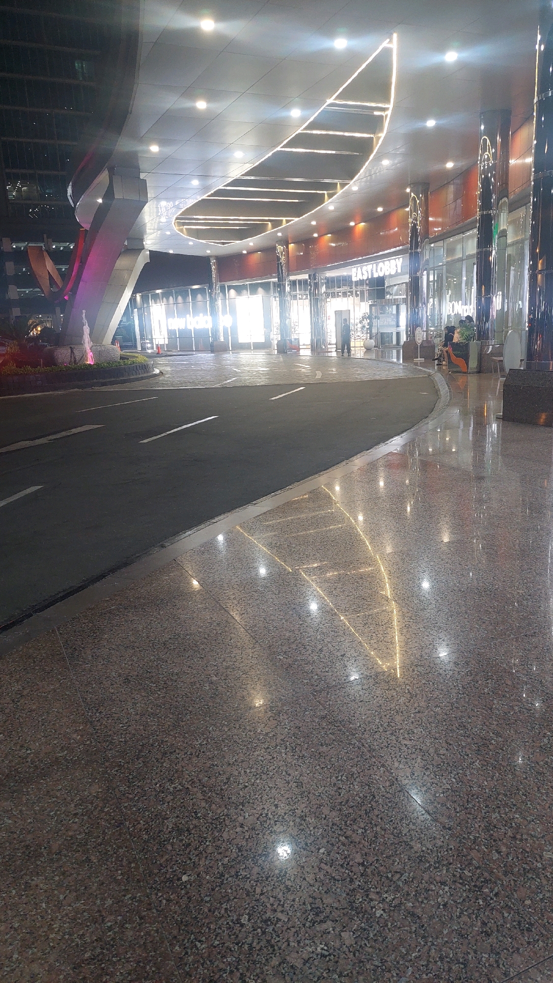 The image depicts the exterior of a modern building at night, likely a commercial establishment or a mall, in Bekasi, Indonesia. The foreground is dominated by a polished, speckled floor that reflects the bright lights of the building's interior and exterior decorative lighting. A dark asphalt road curves into the scene from the left, marked with white lane lines. To the right of the road and extending into the background are the illuminated storefronts of various shops, with one sign clearly visible reading "EAST LOBBY". The architecture features a large, curved overhang supported by pillars. Decorative LED strip lights trace a flowing pattern along the underside of the overhang and inside the lobby area. The scene is lit by a combination of artificial light sources, creating a dynamic interplay of light and reflection on the wet-looking surfaces. There are a few indistinct figures of people visible in the background near the entrances of the establishments, some appearing to be shoppers or employees. A small fountain with pink lighting is visible to the far left, partially obscured by landscaping. The overall atmosphere is one of an active, well-lit urban environment at night.