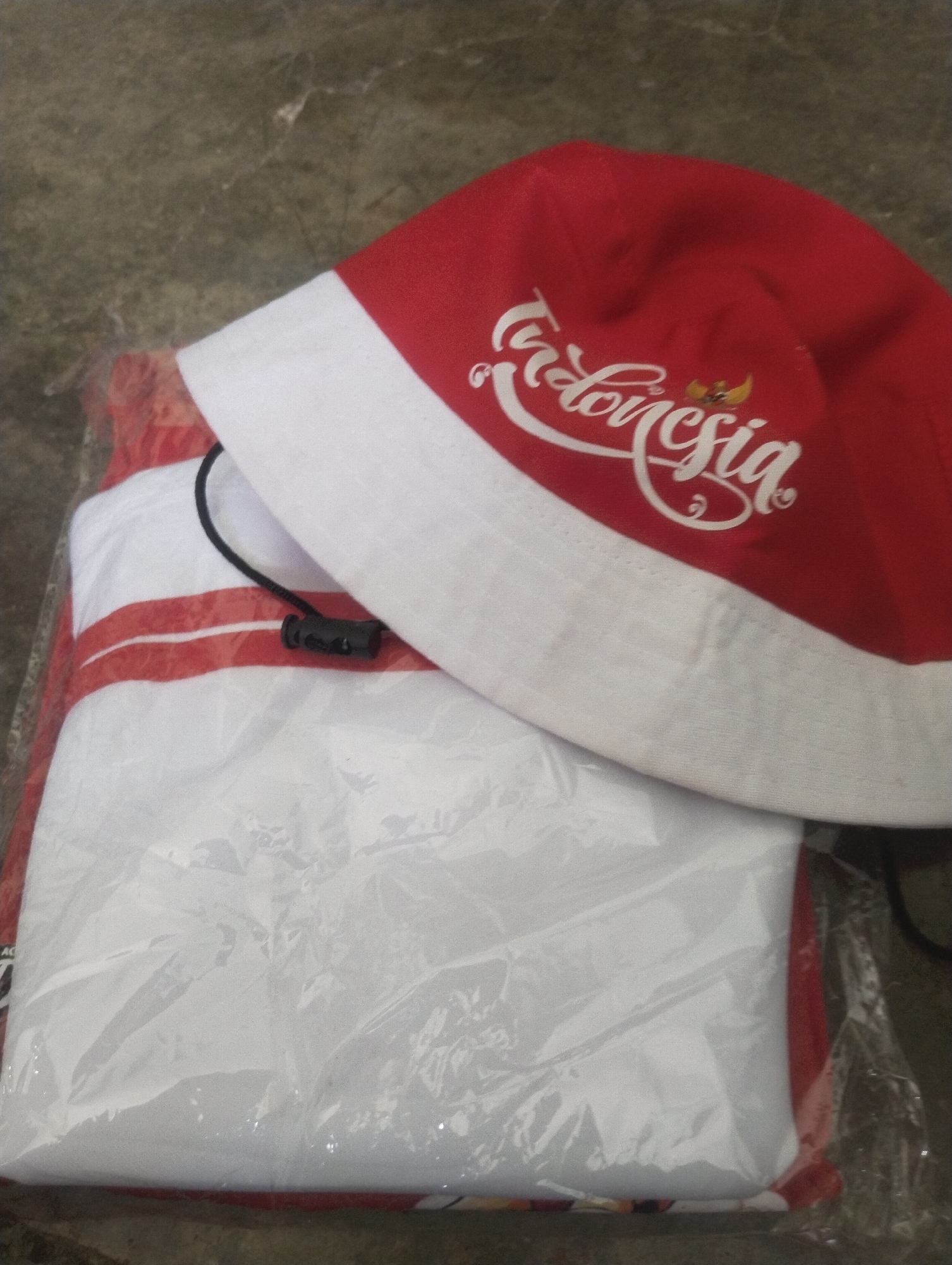 The image shows a red and white bucket hat with the word "Indonesia" written in white cursive script and the Garuda Pancasila symbol. The hat is placed on top of a white t-shirt with red stripes, which is wrapped in clear plastic. The items are on a mottled gray surface, possibly concrete. The location is not identifiable as Wanasaba Daya, Indonesia from the image provided.