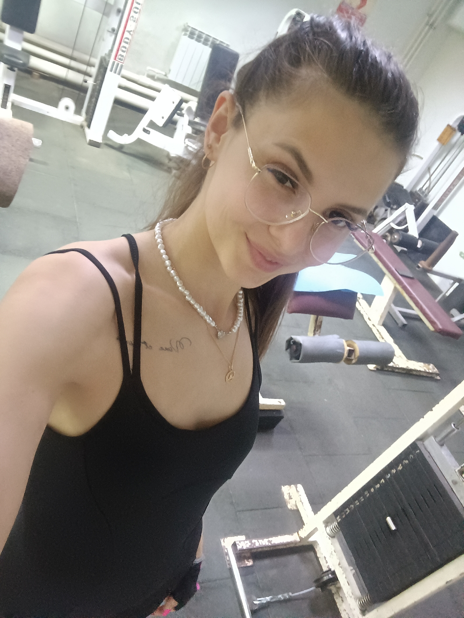 The image shows a woman indoors at a gymnasium. She is fair-skinned, has brown hair pulled back, and is wearing wire-frame glasses. She wears a black tank top, a string of pearls, and a delicate gold pendant necklace. A small tattoo is visible on her upper chest. She is holding the camera, suggesting she is taking a selfie.
Gym equipment surrounds her, including weight machines with weights visible. The gym has white and gray equipment, and the floor is a gray-green color. The location is unknown.