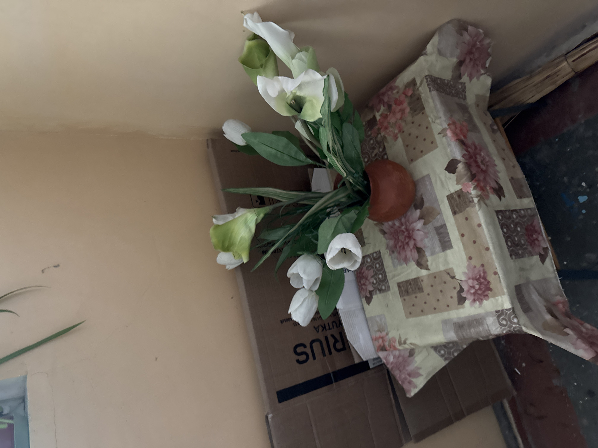 A top-down, angled shot captures a bouquet of white and green calla lilies in a brown vase. The flowers are arranged in a bunch, with some showing their open trumpet-like shapes and others still tightly closed. The backdrop is a light beige wall and a patterned tablecloth with floral motifs and polka dots. The tablecloth is draped over a cardboard box. A broom with a light-colored handle is visible in the upper right corner, leaning against something. The overall lighting suggests an indoor setting with natural light. There is no discernible activity taking place; the scene is a still life composition. Text is visible on the cardboard box, partially obscured, reading "RIUS" and other unreadable characters. The image is rotated approximately 90 degrees counter-clockwise.