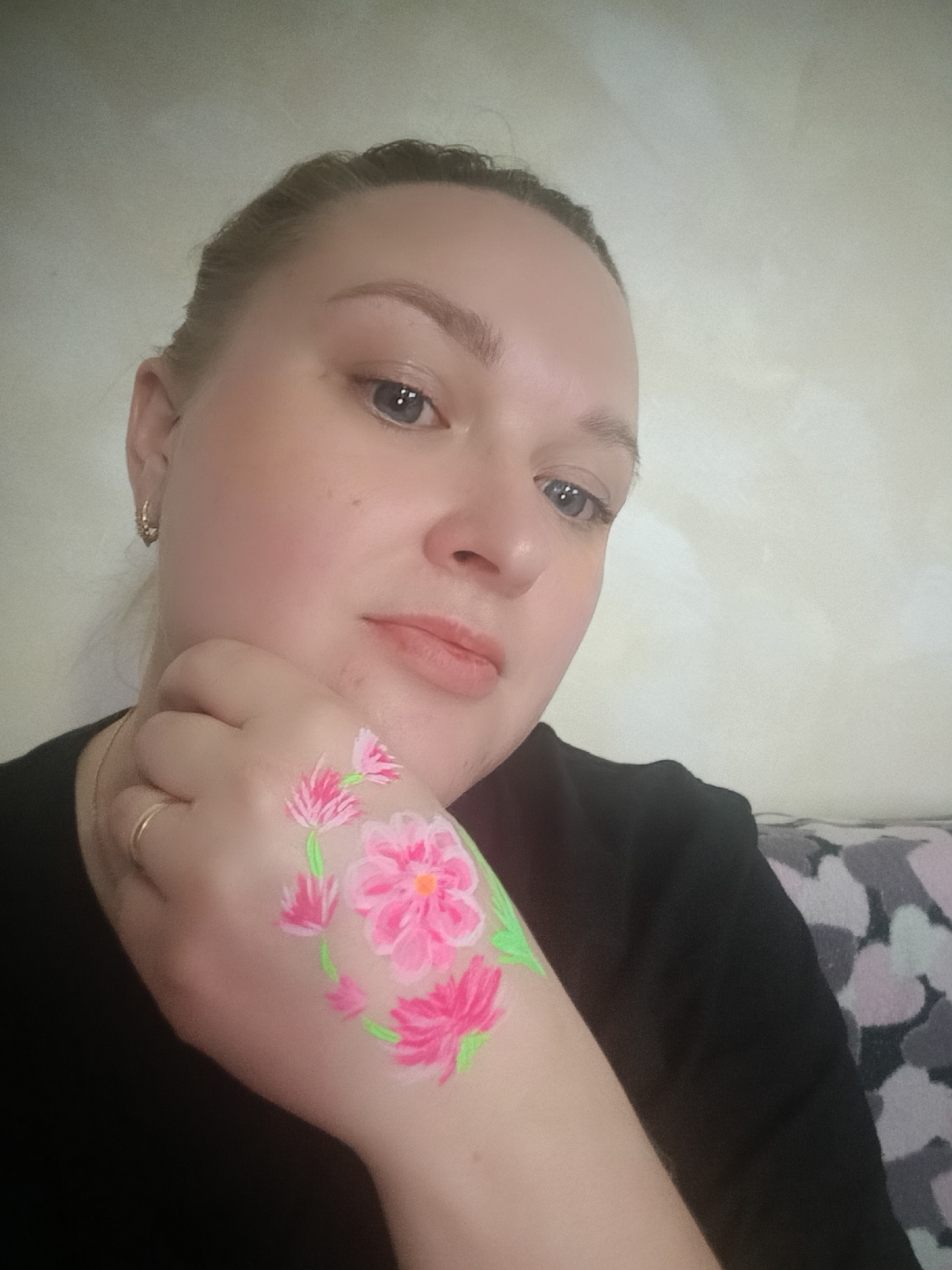 The image features a woman, likely in her late 20s or 30s, posing indoors in Chaikovsky, Russia. She has fair skin, light-colored eyes (appearing blue or grey), and her hair is pulled back. She wears a simple black top.
Her left hand is raised to her face, prominently displaying a vibrant floral body art design on its back. The design consists of several bright pink flowers, some with yellow or orange centers, interconnected by neon green stems and leaves, forming a decorative, wreath-like pattern. She wears a small gold hoop earring in her visible left ear and a simple gold band on a finger of her left hand. Her expression is calm and gentle as she looks towards the camera.
The background consists of a plain, light-colored wall on the left and a patterned cushion or blanket with abstract grey and pink shapes on the right. The lighting is soft and even, typical of an indoor setting. There is no visible text in the image.