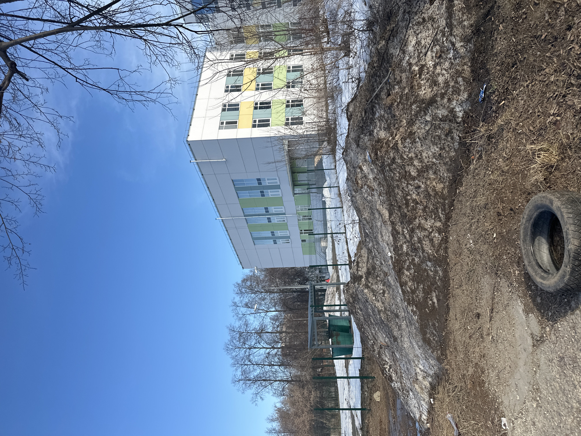 The image shows a partially snow-covered embankment next to a building with alternating yellow and green window panels, likely a school or apartment complex. A discarded tire sits on the ground near the edge of the embankment. The sky is blue with bare tree branches visible. The location is identified as Chaikovsky, Russia.