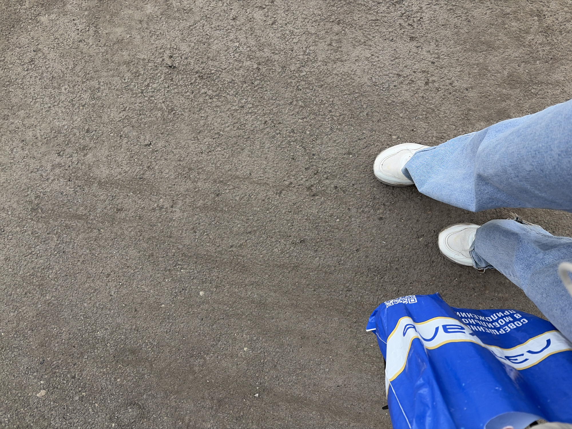 The image depicts an asphalt surface with two legs wearing light blue jeans and white sneakers in the upper-right corner. A crumpled blue plastic bag with white lettering and yellow accents is also visible near the lower-right corner of the image. The lettering on the bag is in Russian and contains the word "AVE". The location is unspecified, but the writing on the bag suggests it could be the city of Izhevsk, Russia.