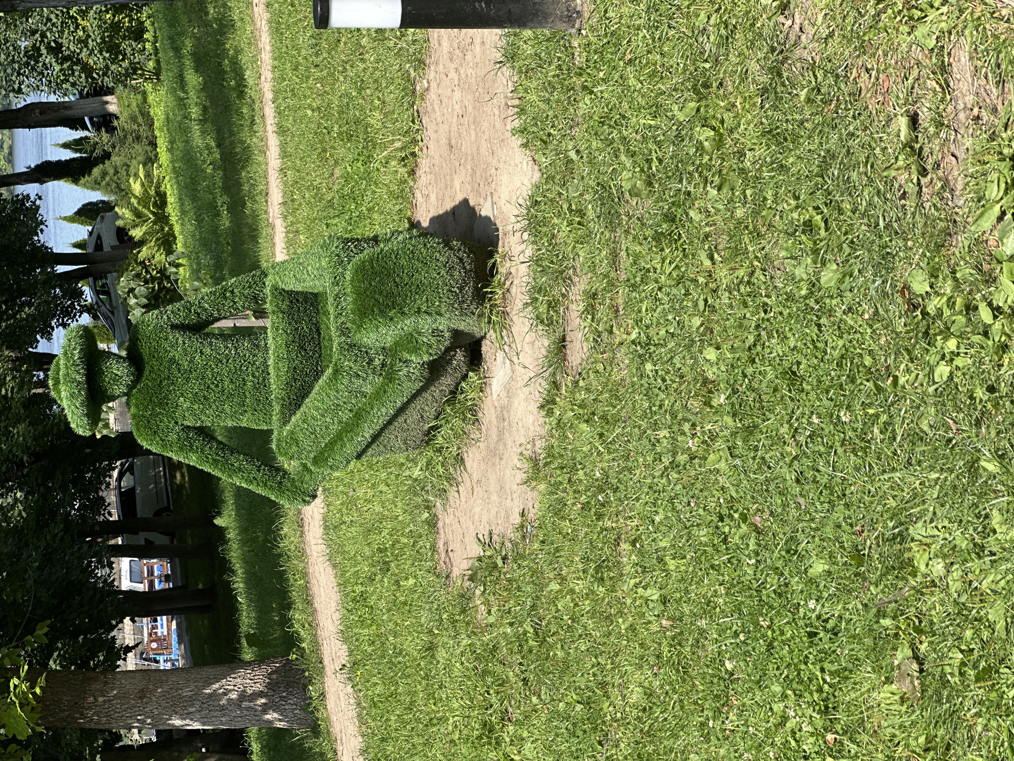 The image shows a topiary sculpture of a person sitting in a wheelbarrow, located in the city of Chelganovo, Russia. The sculpture is made of dense green grass, meticulously shaped to resemble a human figure and a wheelbarrow. The figure is seated with its arms resting on the wheelbarrow, and it appears to be wearing a hat. The wheelbarrow is positioned in front of the figure. The sculpture is situated on a patch of exposed earth surrounded by a gravel path. In the background, lush green grass, trees, and a body of water are visible, suggesting a park or a garden setting. Sunlight casts shadows on the ground. A black and white bollard stands to the right of the sculpture. The overall impression is one of a whimsical and artistic landscape feature in an outdoor public space.