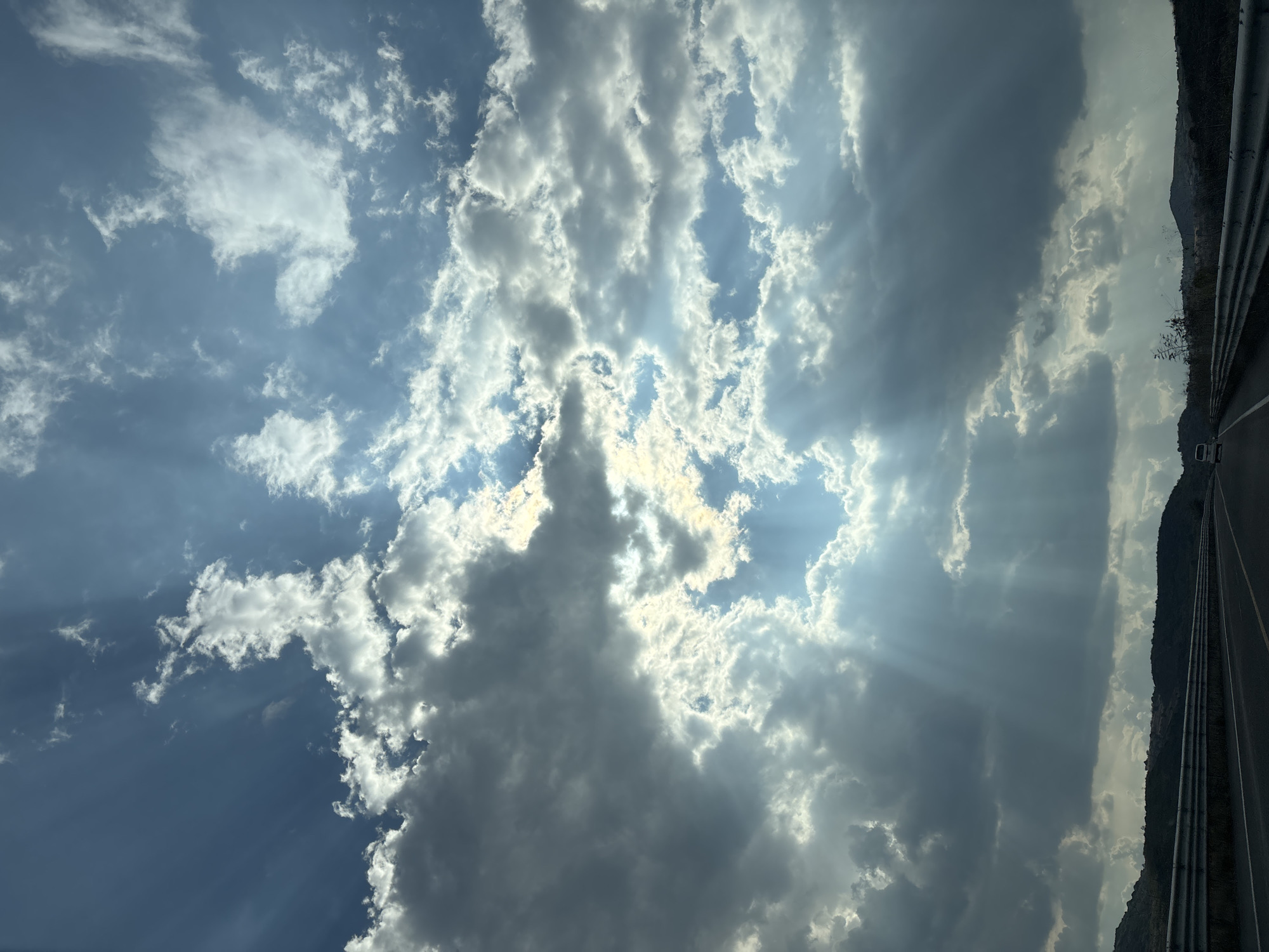 A horizontal, eye-level shot depicts a cloudy sky in what appears to be Santiago Atzitzihuacán, Mexico, though this cannot be definitively verified. The sun streams through a gap in the cloud cover. To the right, a road runs alongside a roadside barrier and an embankment covered with sparse vegetation. There are no people visible in the frame.
