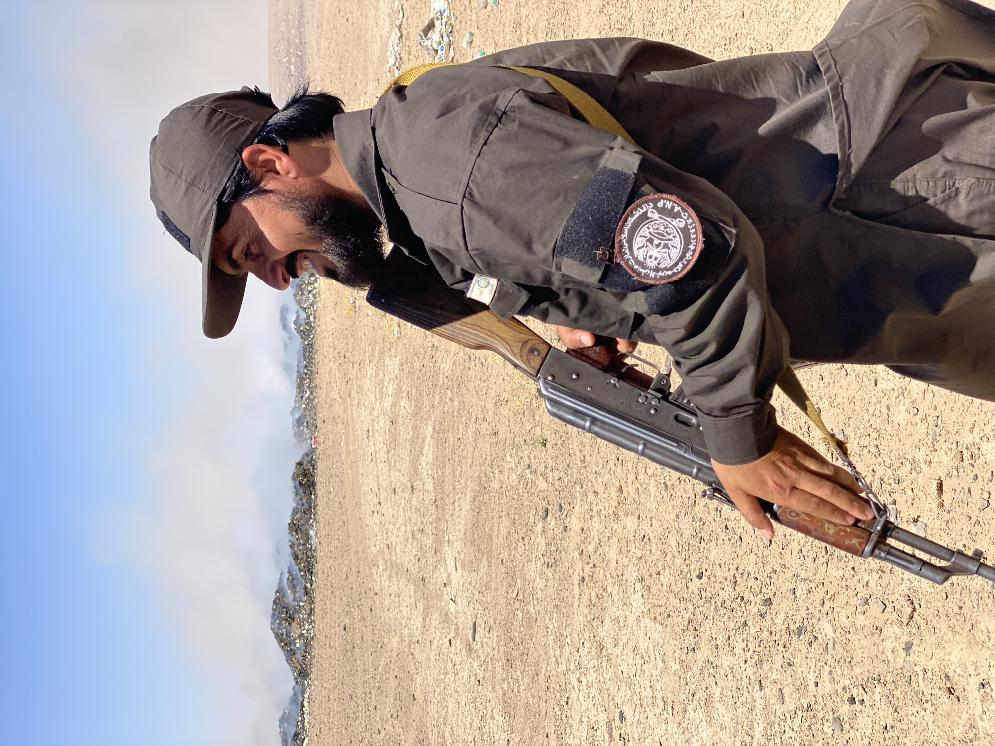 In the city of Mandisar, Afghanistan, a man with a beard and a baseball cap stands in a sandy, arid environment. He is dressed in a dark uniform with patches on his sleeves. He holds an assault rifle, with his hand on the foregrip, and appears to be looking off to his left. Behind him, a large pile of trash is burning, emitting plumes of smoke into the clear blue sky. The ground is covered in sand and small pebbles, with scattered debris visible in the distance. The scene suggests a harsh and possibly neglected area.