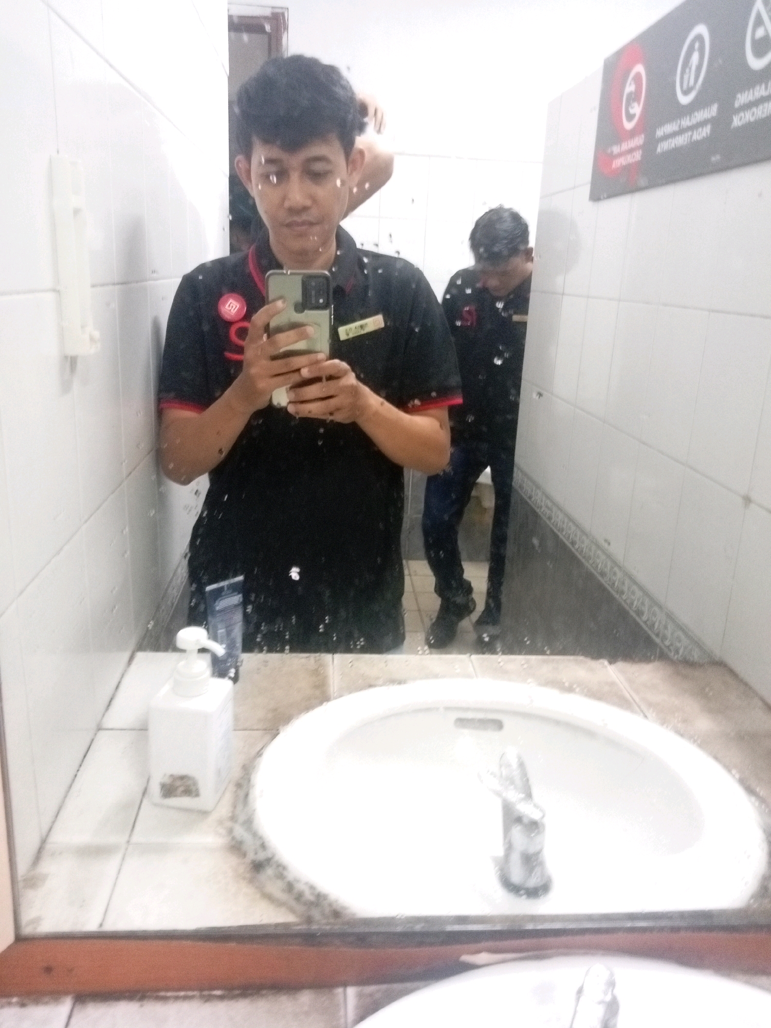A young man in a black polo shirt with red accents and a name tag is taking a selfie in a brightly lit public restroom, likely in Batam, Indonesia. The restroom appears to be tiled with white ceramic, and there are water droplets on the mirror, indicating it is in use. In the foreground, a white sink with a chrome faucet is visible. A white dispenser for liquid soap is also present on the counter next to the sink. In the background, another person is seen bending over, possibly washing their hands or using a urinal, though this is not clearly discernible. A sign with a warning message is mounted on the wall, partially obscured. The overall impression is of a typical, functional public restroom environment.