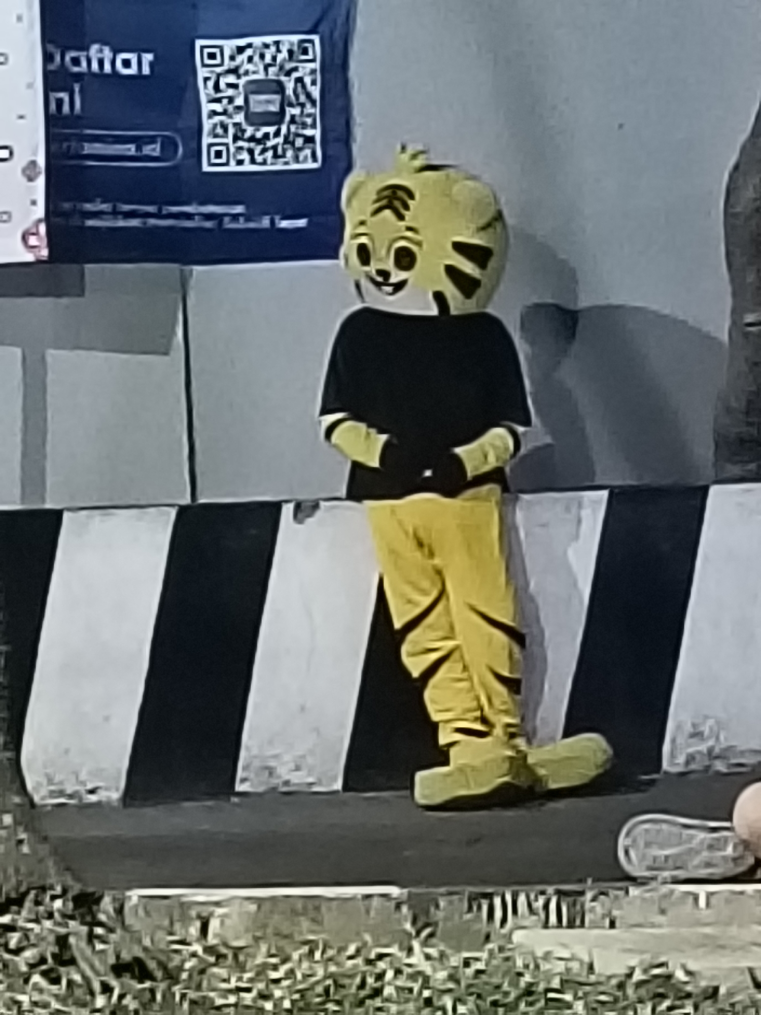 A person in a yellow tiger mascot costume, with black stripes on its head and face, stands with its legs crossed. The mascot is wearing a black shirt and yellow pants, and has oversized yellow shoe-like feet. The mascot is positioned in front of a barrier with alternating black and white diagonal stripes. Behind the mascot, a dark blue banner with white text and a QR code is visible. The environment appears to be outdoors, possibly in an urban setting with a textured ground cover in the foreground. The background consists of a light-colored wall. The image has a low resolution, giving a slightly blurry appearance. The location is identified as Kediri City, Indonesia.