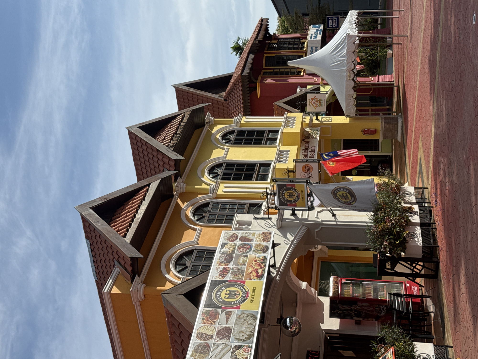 The image captures a row of colorful buildings with distinct architectural styles, set against a bright blue sky with scattered clouds. The buildings are painted in vibrant hues of orange, yellow, and red, with tiled, steeply pitched roofs. The architecture suggests a European influence, with arched windows and decorative moldings.
In the foreground, a red brick-patterned walkway leads up to the buildings. To the left, a section of a restaurant or food establishment is visible, featuring outdoor seating with black chairs and tables. A red Coca-Cola refrigerator is also present. A large banner advertising "German Food Corner" with images of various dishes is displayed prominently on the orange building. A disco ball hangs from an overhang.
Several flags are visible: a red and white flag, and the Malaysian flag. One building has signage that reads "gelato tradisional Kedah". Further to the right, a white canopy tent stands in front of a red building. Another sign indicates "Fish & Chips". The overall scene appears to be an outdoor dining or entertainment area in a tourist-oriented location. The location is likely a food court or a street lined with shops and restaurants. Based on the signage and architecture, it is plausible that this is located in Langkawi, Malaysia, known for its tourist attractions and diverse culinary offerings. There are no people visible in the frame.