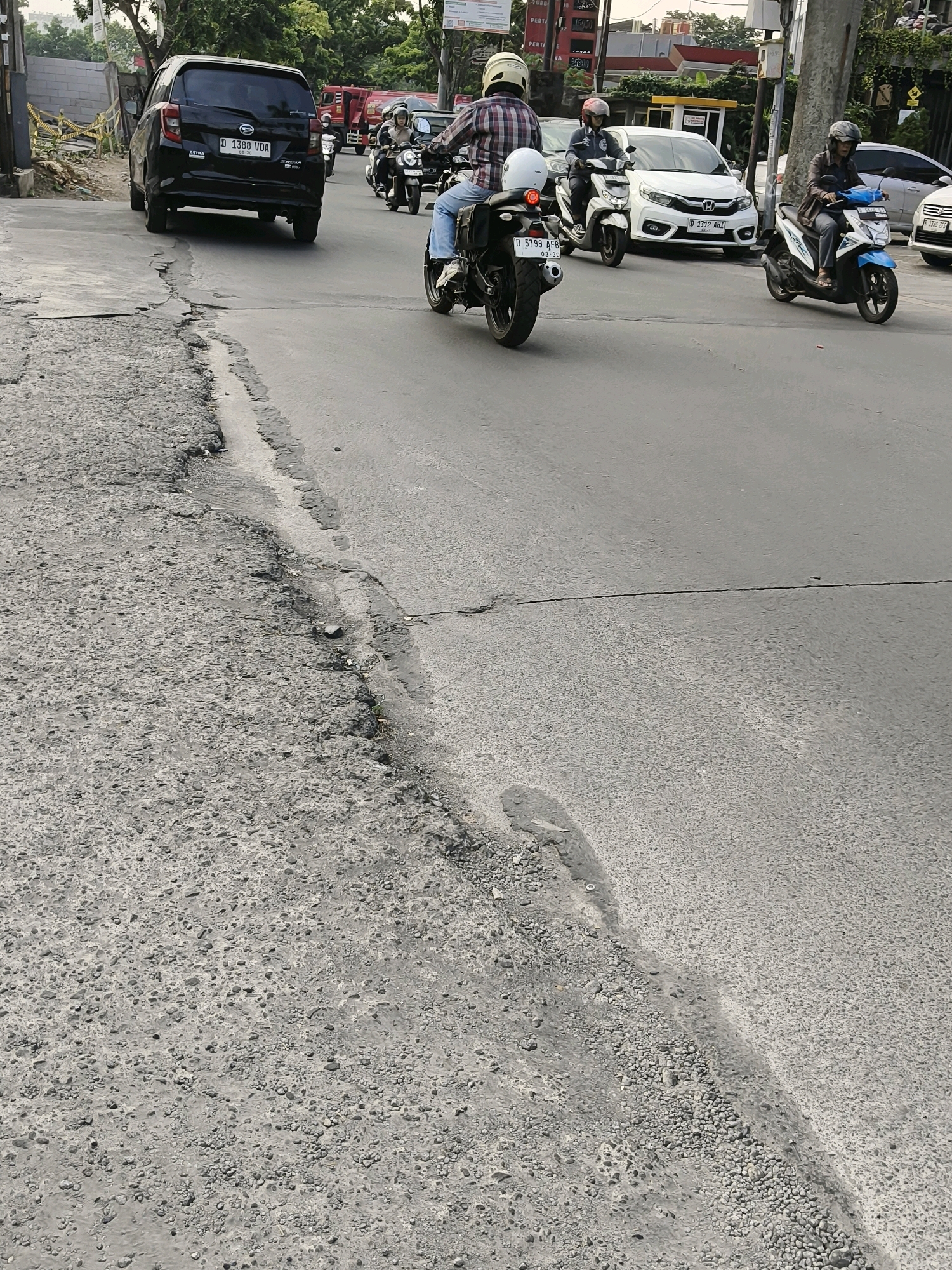 In Bandung City, Indonesia, a road scene unfolds with multiple vehicles and a damaged road surface. A black Daihatsu Sigra with license plate D 1388 VDA is parked on the left. Ahead of it, several motorcycles are in motion. One rider on a black motorcycle, wearing a plaid shirt and a white helmet, is moving away from the camera. Another rider on a white motorcycle, wearing a helmet, is also in motion. Further ahead, a rider on a blue and white motorcycle, wearing a brown jacket and helmet, is traveling on the road. The road itself exhibits significant damage, with a cracked and crumbling asphalt edge on the left side. In the background, fire trucks are visible, suggesting a potential incident or emergency services presence. The environment appears to be a moderately busy urban street.