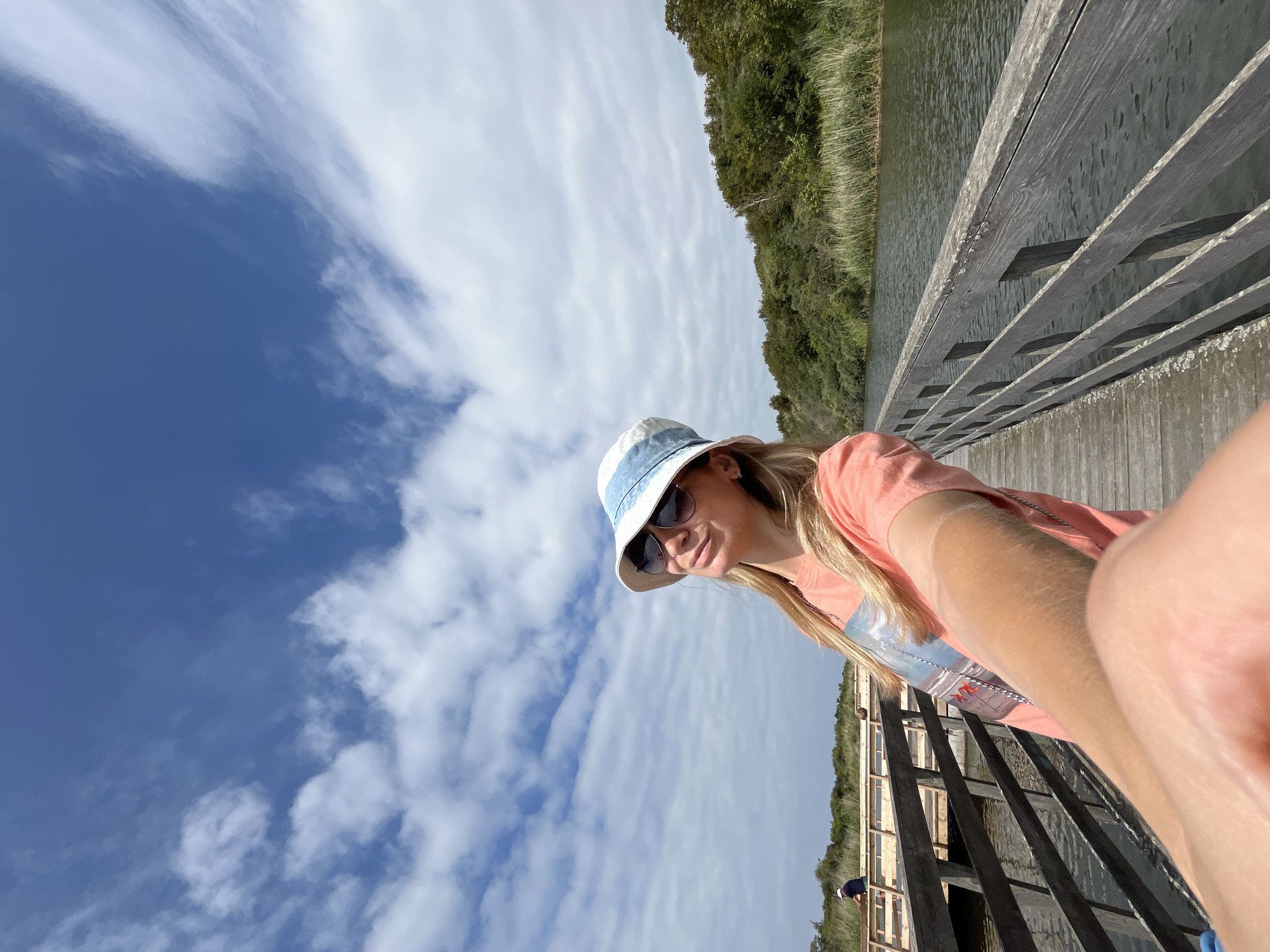 A woman with blonde hair, wearing a peach-colored t-shirt and a white and blue bucket hat, takes a selfie on a wooden boardwalk. She is also wearing sunglasses. The boardwalk spans over calm water and is flanked by a railing. In the background, there is lush greenery and trees, with a bright blue sky dotted with white clouds overhead. A small section of a different boardwalk is visible in the distance to the left, where a person wearing a dark blue shirt and a white cap is seen tending to something. The scene appears to be a natural environment, possibly a park or reserve, located in Yantarny, Russia.