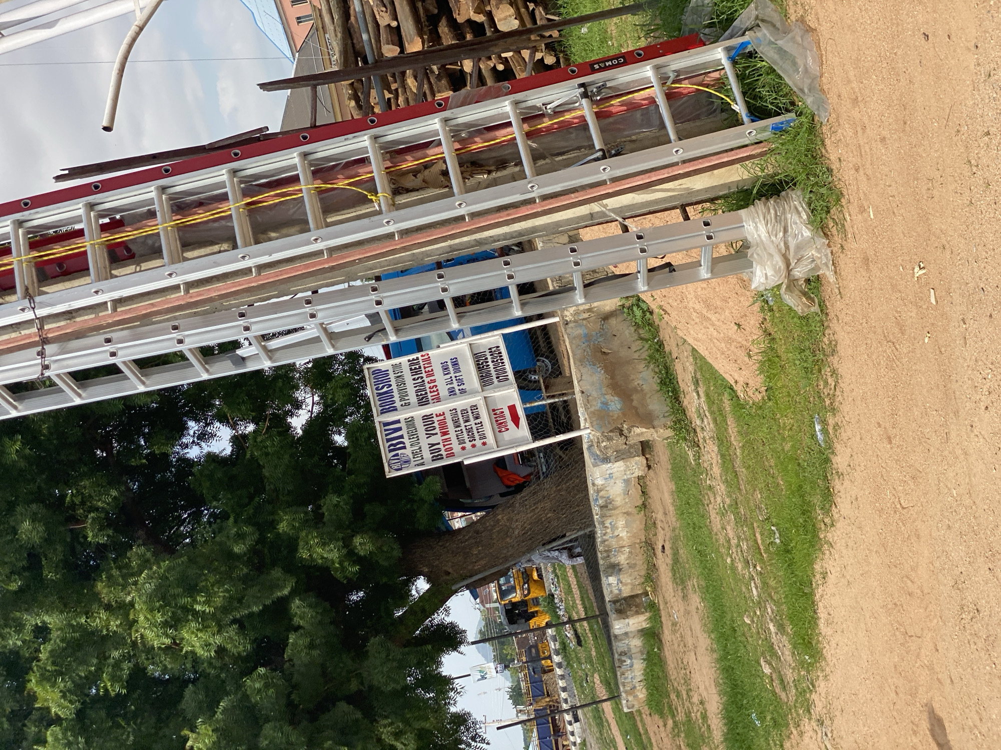 This media file depicts an outdoor scene in Jalingo, Nigeria. The primary objects in focus are several aluminum ladders stacked together. One ladder, with red accents, is prominently featured, while another silver ladder is positioned below it. The ladders appear to be new or in good condition.
In the background, there are piles of cut wood, suggesting construction or a woodworking area. Behind the wood, a building with a pink facade and a blue tarp is visible. To the left, a lush green tree with dense foliage dominates the frame.
A signpost is also clearly visible. It displays "BIVI BOOKSHOP" and lists various items for sale, including "LEVEL/OLEVEL BOOKS & PROVISION STORE," "MINERALS," "BOTTLE MINERALS," "SACHET WATER," "BOTTLE WATER," and "SOFT DRINKS." Contact numbers are also listed on the sign.
The ground is a mixture of dry, sandy earth and patches of green grass. In the distance, several yellow vehicles, possibly buses or trucks, are parked or in motion, indicating a public area or a roadside setting. The sky is partly cloudy, suggesting daylight. The overall impression is one of a commercial or industrial area, possibly a marketplace or a construction site, with a focus on the ladders and the bookshop. There are no people clearly visible in the image. The scene is brightly lit, indicating it was taken during the day, likely in the morning or afternoon.