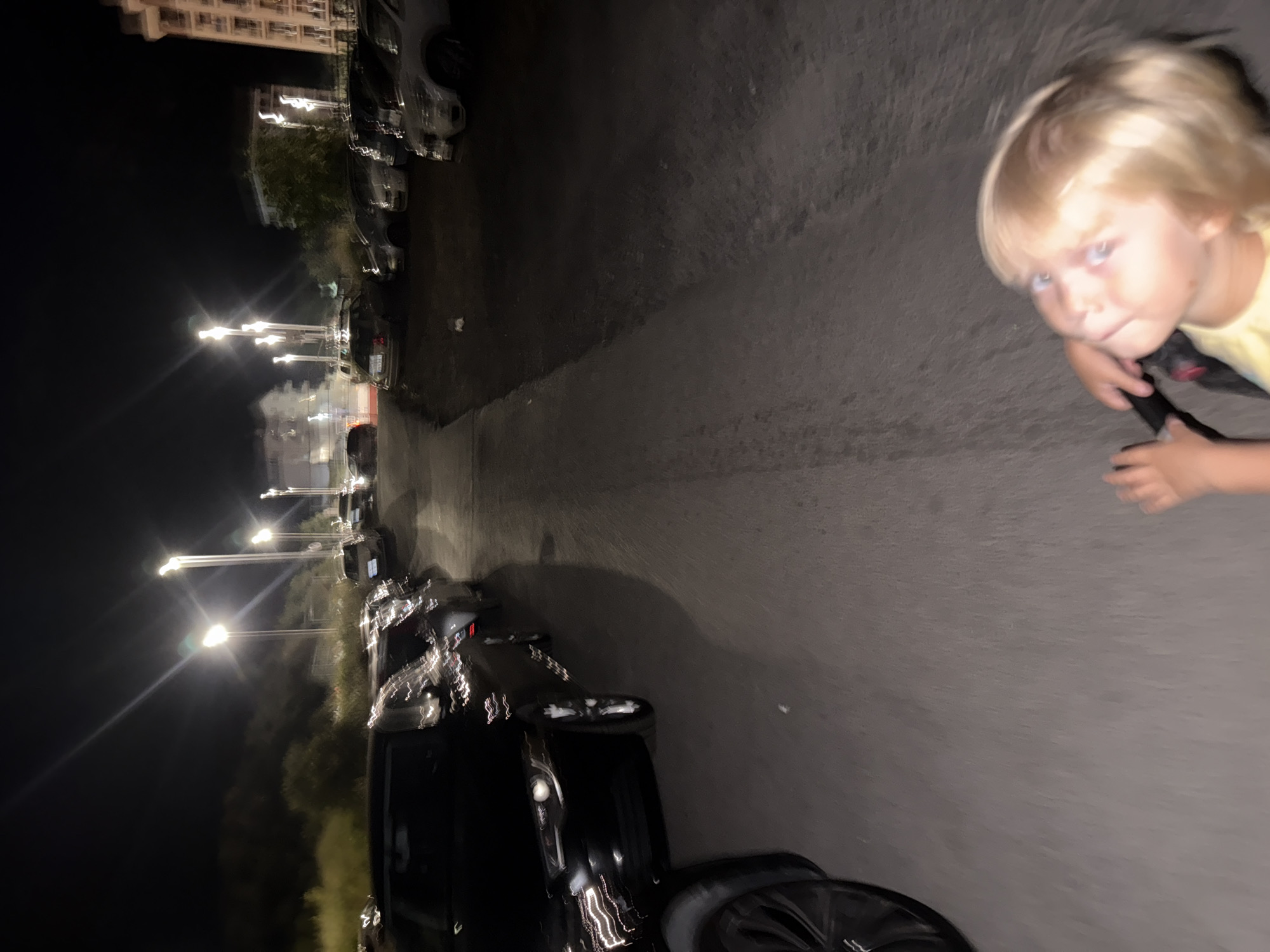 In the city of Boreti, Montenegro, at night, a young child with blonde hair is looking upwards. The child is holding onto what appears to be the handlebars of a stroller or bicycle. In the background, numerous cars are parked on the side of a street. Streetlights illuminate the scene, casting long shadows and creating lens flares due to the low light conditions. Buildings and trees are also visible in the distance. The image is blurry, suggesting movement during the capture.