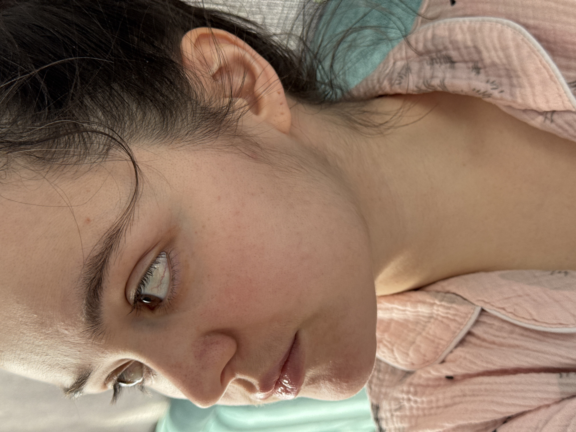A close-up, angled view depicts a person with dark hair, light skin, and brown eyes. The individual is wearing a pink pajama top with a light pattern. The background consists of a pale green or blue-green surface. No information regarding the city of Izhevsk, Russia is present in this image.