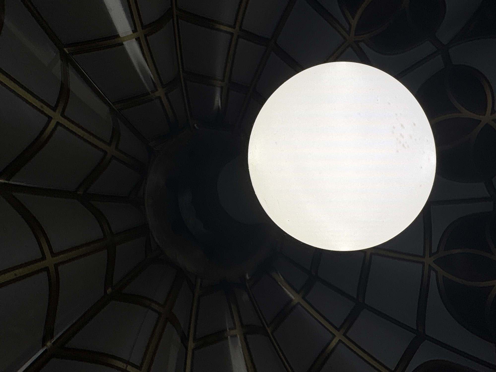 The image shows a close-up, downward-facing view of a light fixture within a dark, structured environment. A bright, circular light source dominates the lower half of the frame. Above and behind it, a darker, curved surface with a patterned grid of intersecting dark lines and golden accents is visible. The pattern suggests a domed or vaulted ceiling. The overall impression is of an interior space, possibly a modern building or a decorative ceiling installation. The provided hints do not offer any specific details about the location in the city of Dnipro, Ukraine, or any discernible context beyond the visual elements of the light fixture and its surrounding structure.