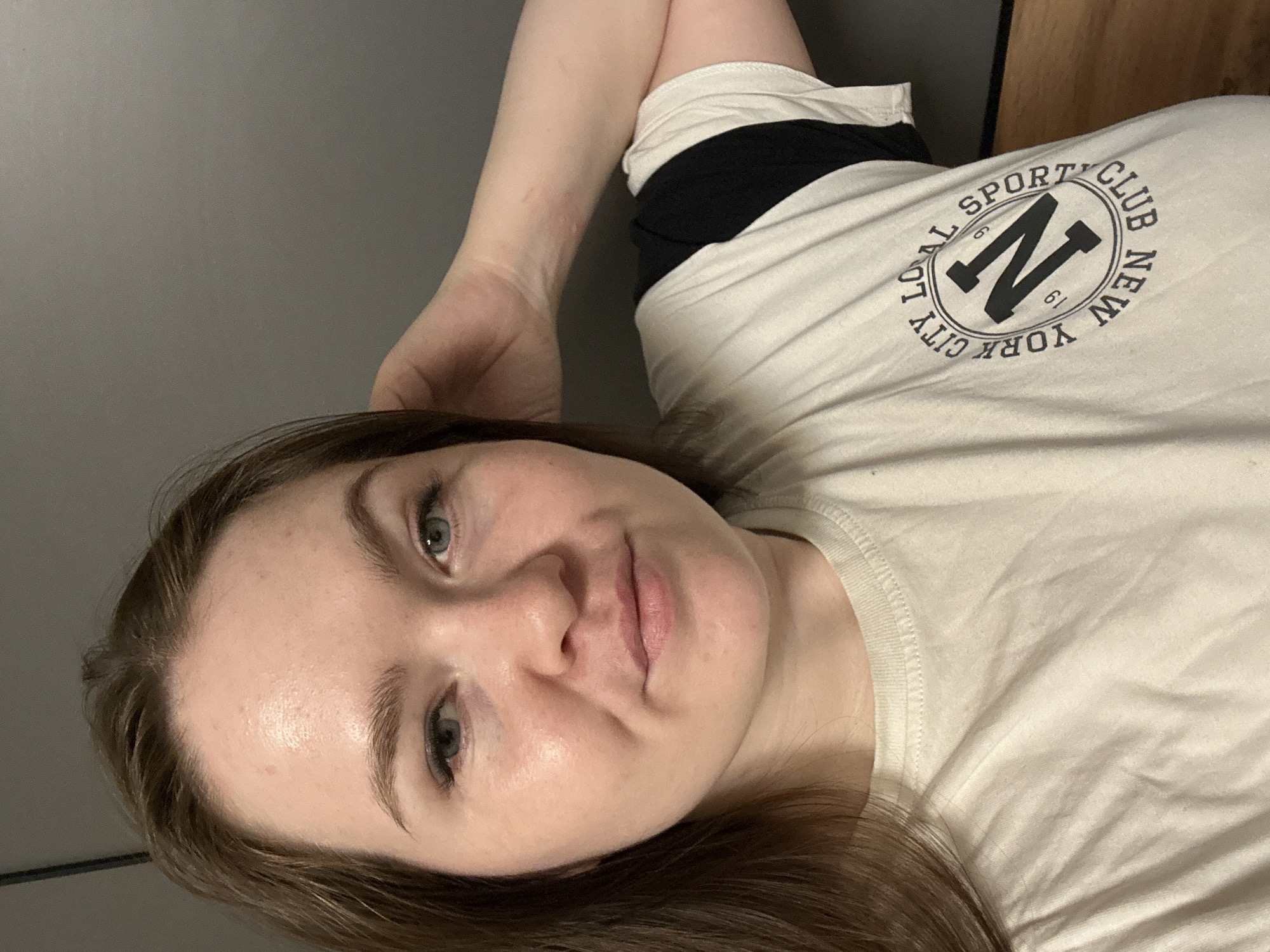 A selfie of a fair-skinned woman with brown hair and light eyes wearing a cream-colored t-shirt with a black graphic design reading "Local Sports Club New York City 1961" with a large "N" in the center. The woman is reclining, with her right arm bent and her hand near her head. The background is a plain gray wall and a partial view of a wooden surface. There is no visible connection to the city of Chaikovsky, Russia.