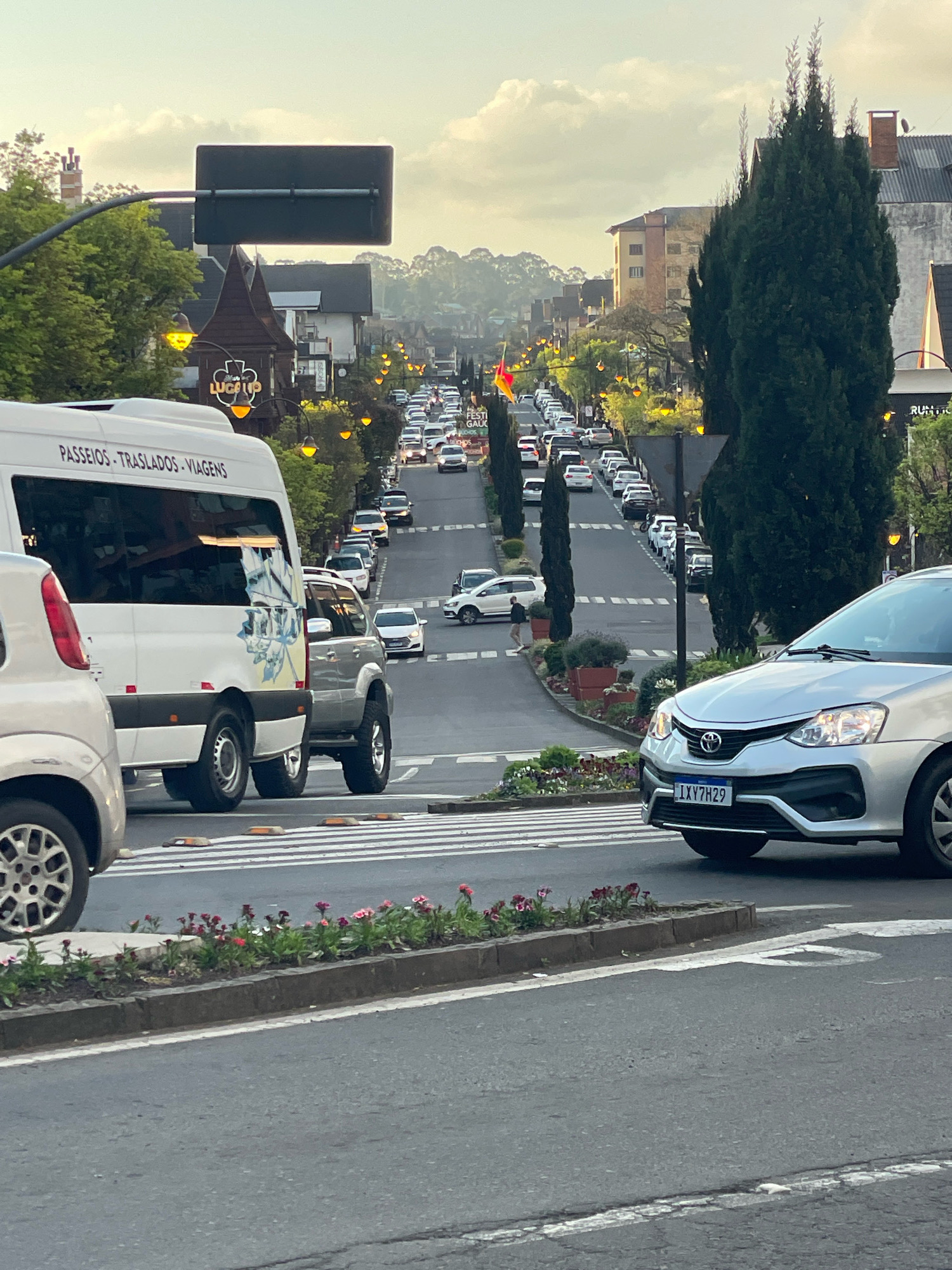 The image depicts a street scene in Gramado, Brazil, likely taken from a slightly elevated perspective. The street is lined with trees and buildings, many of which have a European architectural style, common in Gramado's downtown area. A long line of cars is visible moving up a hill, suggesting moderate traffic congestion. Streetlights with a distinctive design are illuminated along the road, indicating it is either dusk or a cloudy day.
In the foreground, several vehicles are present. A white van with text that appears to say "PASSEIOS - TRASLADOS - VIAGENS" (tours - transfers - trips) is on the left, indicating it is likely a commercial transport vehicle. To its right is a silver sedan with a Brazilian license plate "IXY7H29". The vehicles are positioned on a road with a pedestrian crossing clearly marked by white stripes. On the right side of the road, a large coniferous tree stands tall.
The environment appears to be a commercial or tourist area, with businesses suggested by the building facades and signage. The overall atmosphere is peaceful, with the soft lighting and the gentle flow of traffic. The presence of decorative planters with flowers adds to the aesthetic appeal of the street. The sky is overcast with some hints of sunlight peeking through.