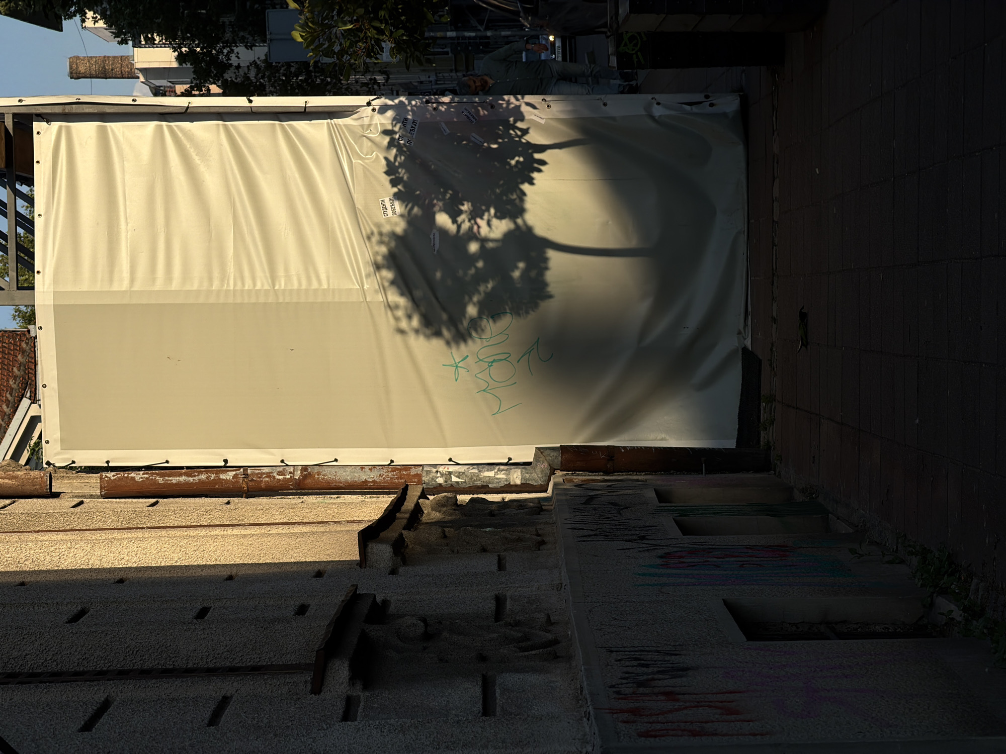 A view of a construction site in Belgrade, Serbia, from street level. A white canvas cover is affixed to scaffolding to obscure the site. The shadow of a tree is cast onto the canvas. Graffiti is visible on the canvas and on the concrete structures in the foreground. The structures consist of exposed and broken concrete steps or platforms. A dark brick or concrete wall is visible on the right side of the frame. Vegetation grows near the wall. The overall lighting suggests an outdoor environment with the sun casting harsh shadows.