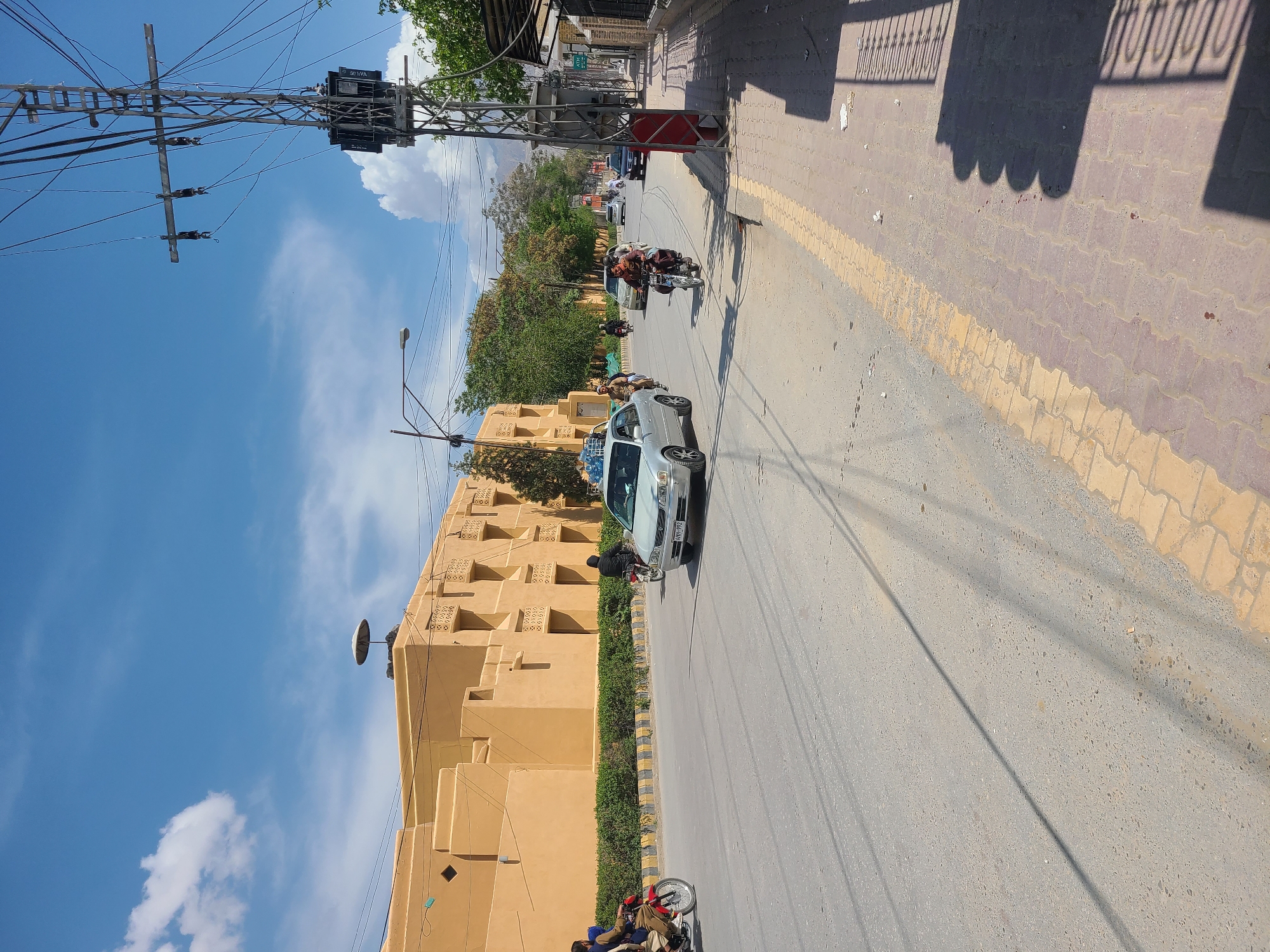 The image shows a street in Quetta City Tehsil, Pakistan. A light gray car is driving on the road, and two motorcycles are also visible. The sky is blue with some clouds. A large, orange building is on the left side of the road, and power lines run overhead. The road surface is asphalt, and there is a yellow line marking the edge of the street. Pedestrians are also present on the road.