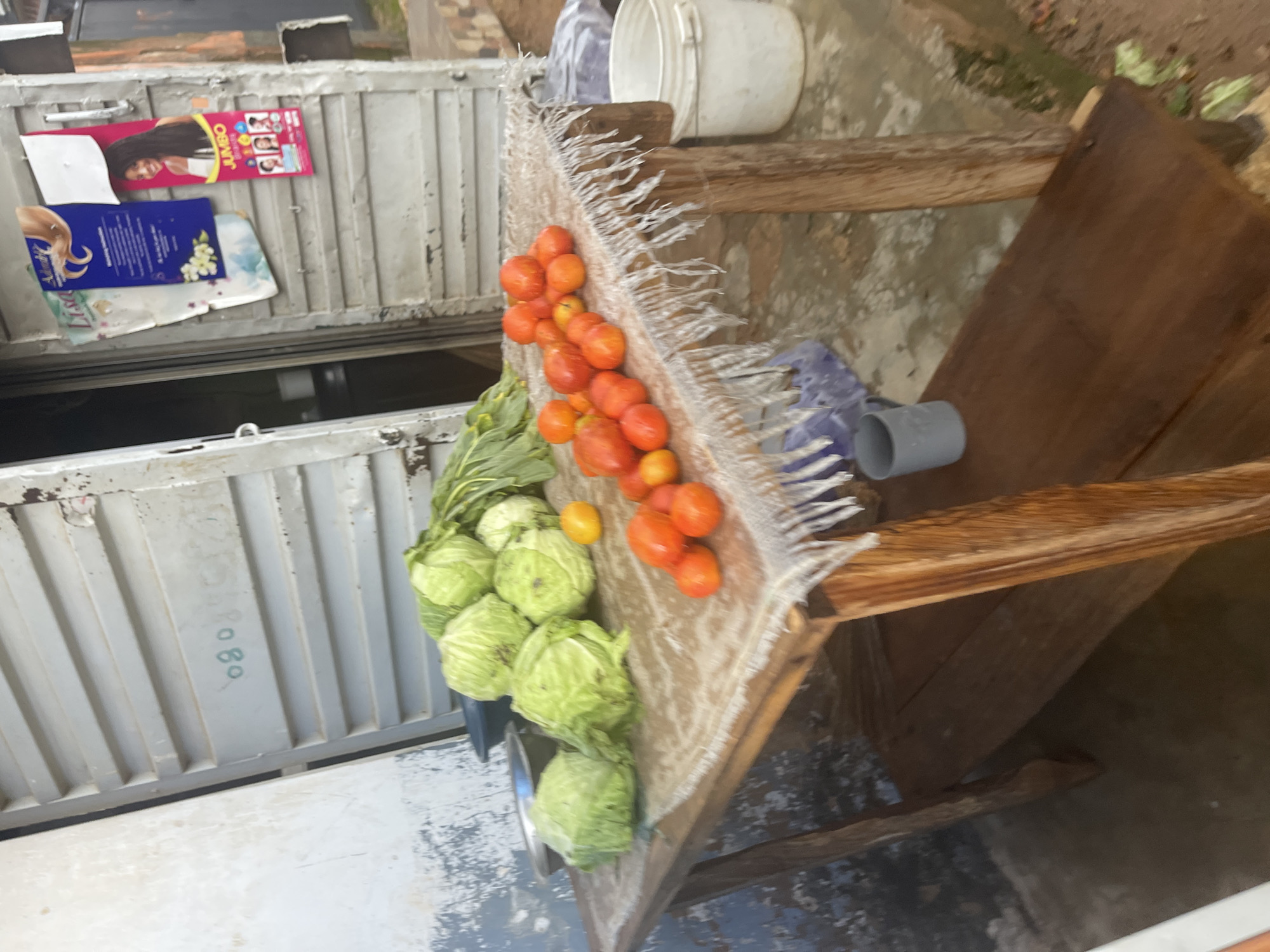 A wooden table with a fringed cloth is laden with fresh produce, including several heads of cabbage and a pile of ripe tomatoes. The scene is set against the backdrop of a rustic building with corrugated metal doors, one of which is adorned with advertisements. A white bucket and a gray mug are visible near the table. The ground appears to be a dirt or concrete surface, suggesting an outdoor market or roadside stall, likely in Jos, Nigeria. The overall impression is that of a simple, vibrant scene of local commerce.