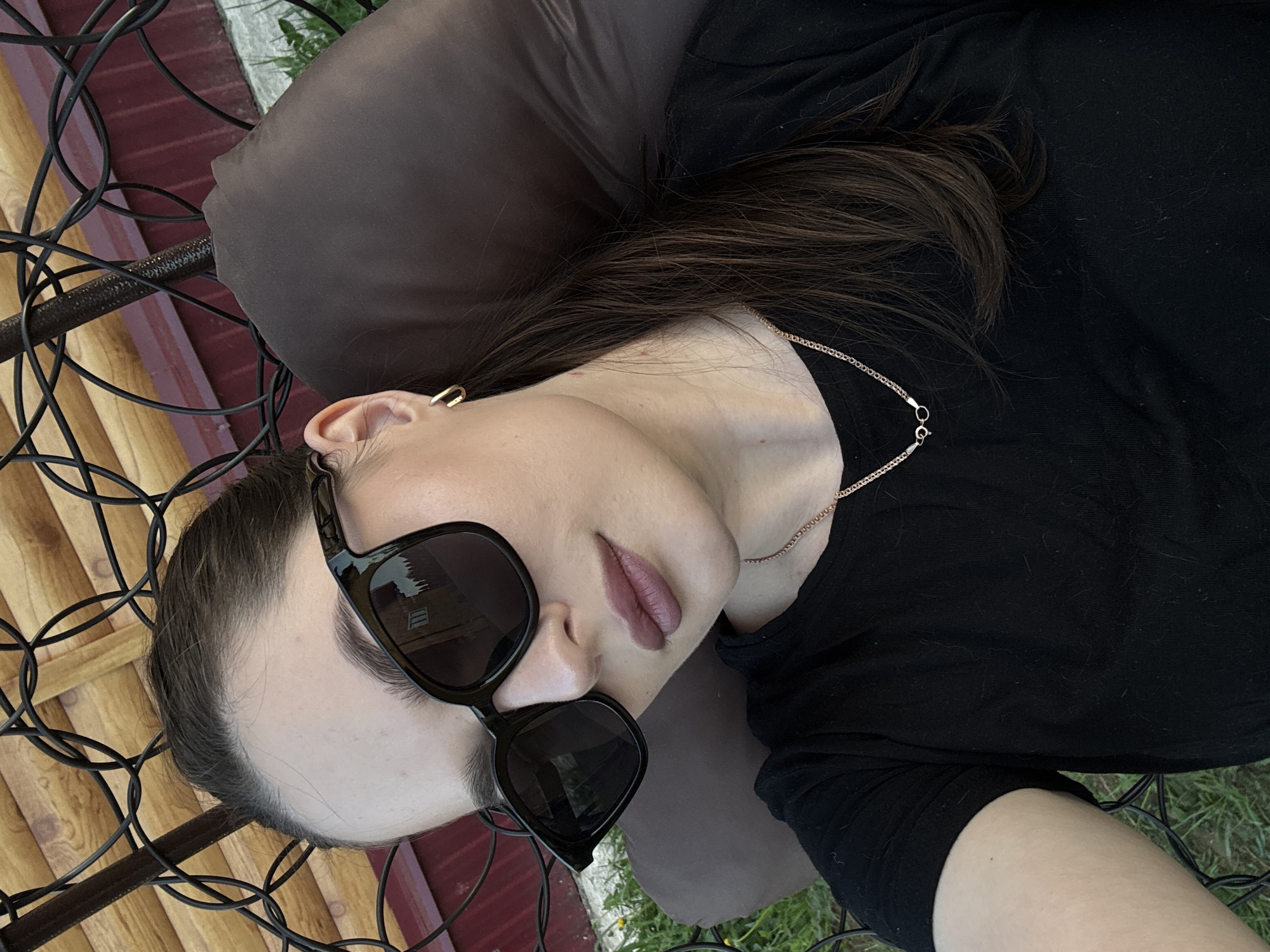 A woman is lying down on a chair or lounge. She is wearing sunglasses and a black top. A brown pillow supports her head. Part of a wooden structure is visible in the background, and there is green grass in the lower right corner of the image. A gold chain is visible around her neck. The location is unspecified beyond the image itself. The image does not provide enough information to confirm it was taken in Rusalevka, Russia.