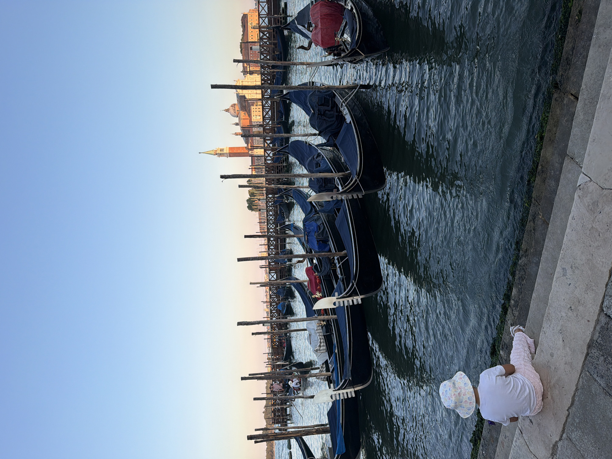 The image depicts a serene waterfront scene in Venice, Italy, likely during the late afternoon or early evening. In the foreground, a young child wearing a white t-shirt, pink patterned pants, and a white bucket hat sits on a concrete ledge, facing away from the camera and towards the water. The child's gaze is directed at a row of moored gondolas.
The water is calm, reflecting the clear blue sky and the surrounding structures. A line of dark-colored gondolas, each with its characteristic pointed prow and oar, is docked along the waterfront, secured by wooden poles. Some gondolas are covered with tarps, while others are partially exposed. In the background, a distant view of Venice's iconic architecture is visible, including a prominent bell tower and domed buildings, suggesting a location near St. Mark's Square or the island of San Giorgio Maggiore. A wooden walkway or pier runs parallel to the gondolas, with a few people visible in the distance, including one person in a gondola being rowed. The overall atmosphere is tranquil and picturesque, characteristic of Venice's unique charm.