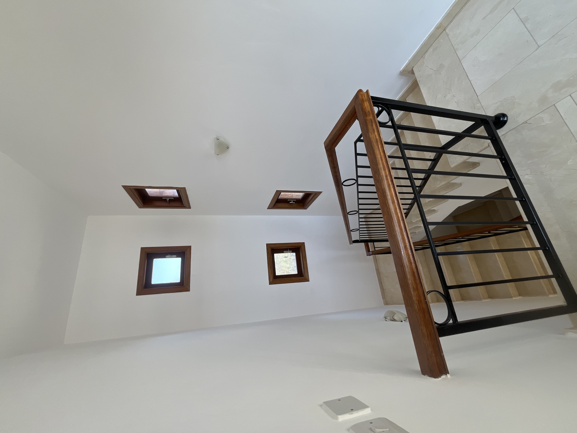 The image depicts an interior staircase area within a modern residential property at Aphrodite Hills, Cyprus. The walls are painted white, and the flooring is a light-colored marble tile. A wooden handrail with black metal balusters lines the staircase, which is visible descending. The staircase features light-colored marble steps. On the walls, there are several small, square windows with wooden frames, some of which offer a view of the sky and greenery. A wall-mounted light fixture is visible on the upper right wall. Electrical outlets and light switches are present on the left wall. The scene is well-lit, suggesting daytime.
