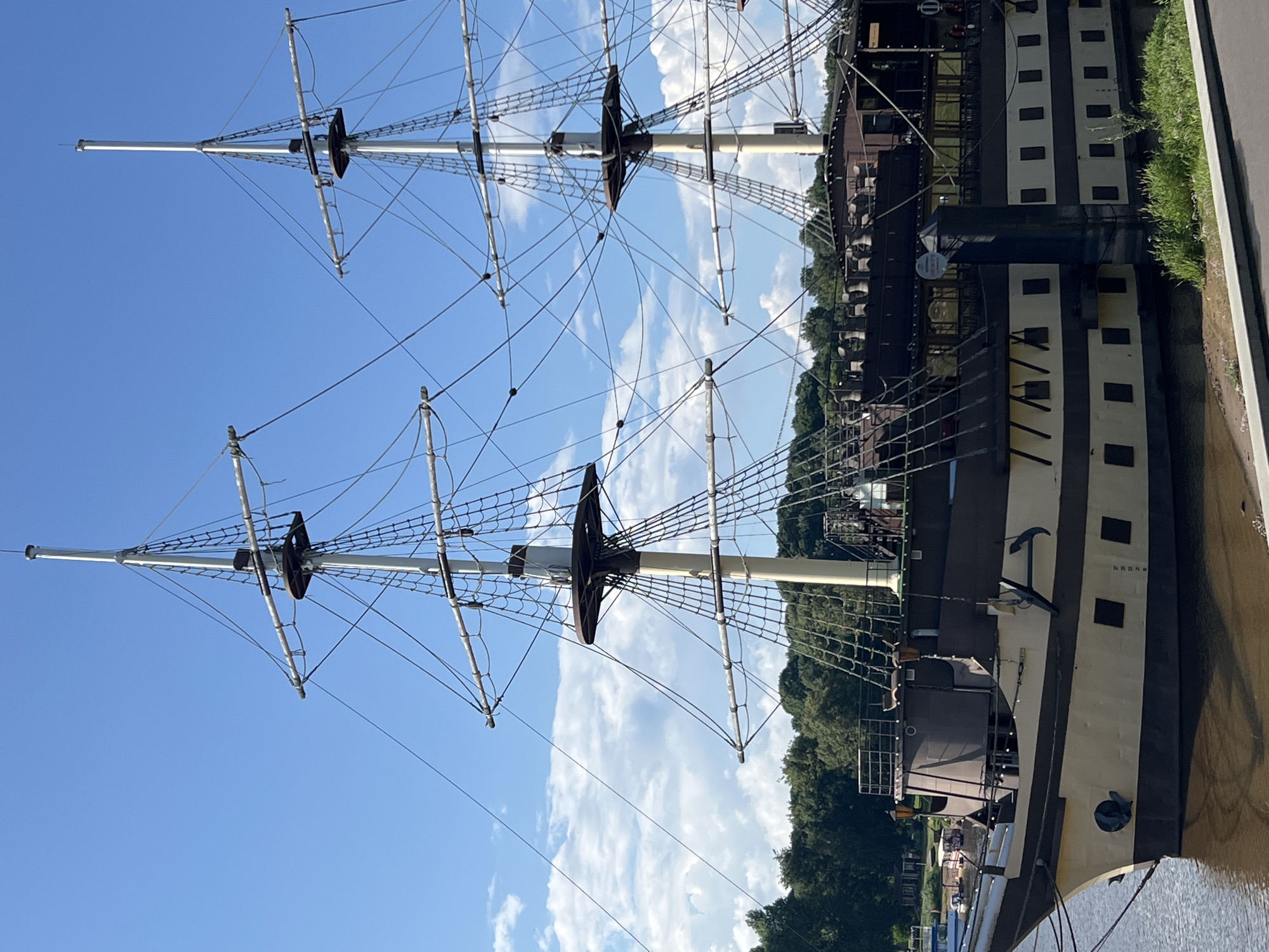 The image shows a large, multi-masted sailing ship docked on the Volkhov River in Veliky Novgorod, Russia. The ship has a distinct tan and dark brown hull, with multiple decks and windows visible. Several masts with rigging are prominent against a bright blue sky with scattered white clouds. Trees and vegetation line the far bank of the river. The ship appears to be a tourist attraction or restaurant, given its elaborate design and the presence of docked smaller boats in the background. There are no people visible in the image. The water is calm, reflecting the ship and the sky. The foreground shows a paved pathway and some grassy embankment.