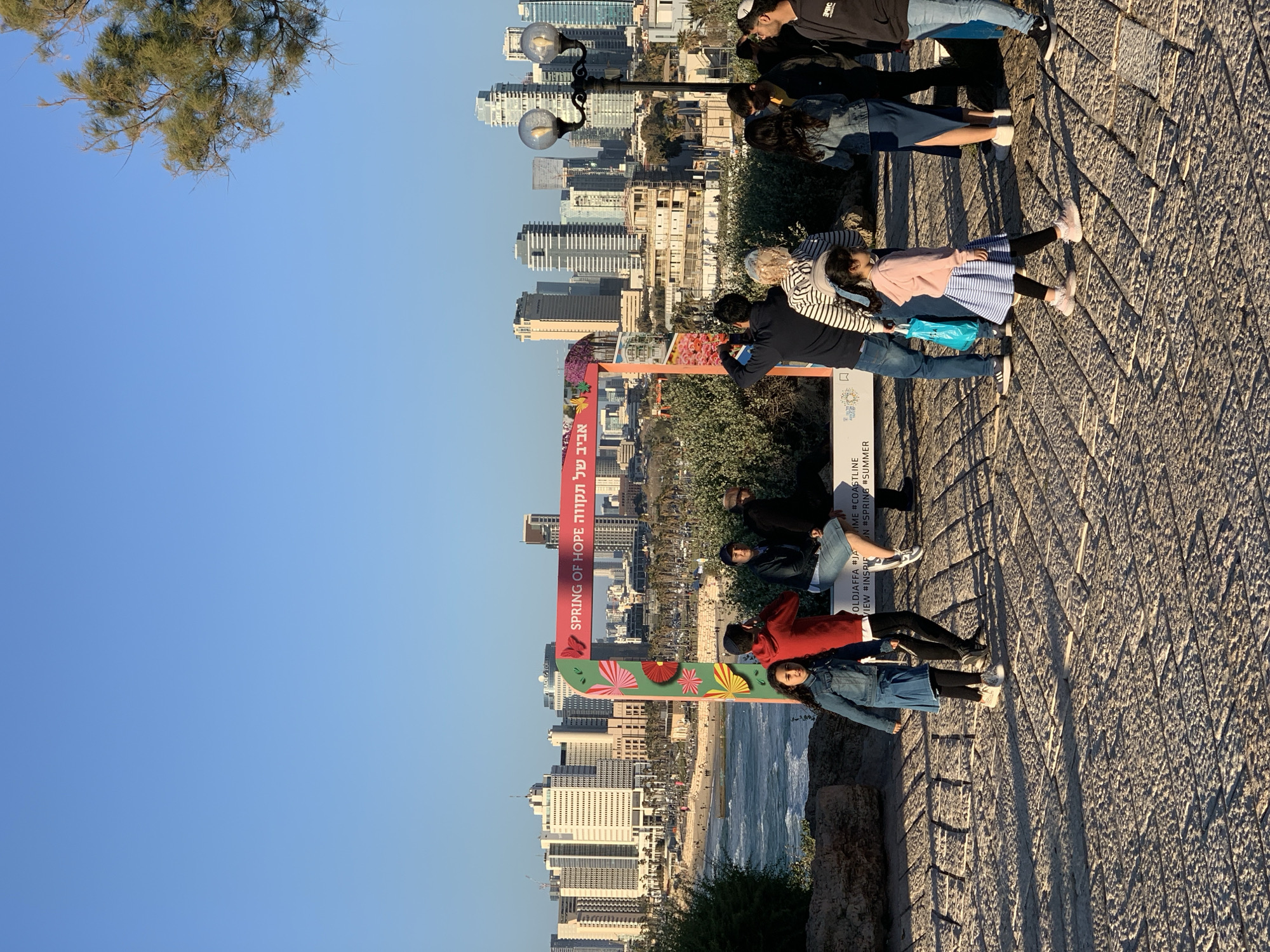 The image presents an outdoor scene from an elevated promenade in Tel-Aviv, Israel, offering a panoramic view of a coastal city and the Mediterranean Sea under a clear blue sky. The warm, directional lighting and long shadows suggest either morning or late afternoon.
Several individuals, including adults and children, are gathered on the textured, paved ground of the promenade. They appear to be observing the scenic vista. One adult is seen holding a small child, while others stand near a prominent, decorative red-framed sign that faces the city view. This sign is adorned with colorful fan-like or floral illustrations.
Beyond the promenade, a sandy beach stretches along the coast, meeting the sea where gentle waves are visible. The backdrop is dominated by a dense urban skyline, featuring numerous modern high-rise buildings and skyscrapers lining the coastline. Two classic black lampposts are also visible on the promenade, and a tree branch with green foliage extends into the upper left corner of the frame.
The visible text on the red sign includes "אביב של תקווה SPRING OF HOPE" in both Hebrew and English. Further text on a smaller panel provides contextual clues such as "OLDJAFFA #JA", "VIEW #INSPIF", "IME #COASTLINE", and hashtags like "#SPRING" and "#SUMMER", confirming the location as Old Jaffa or a viewpoint overlooking it, and referencing the seasonal appeal of the area. The overall activity involves enjoying the picturesque urban coastal landscape.