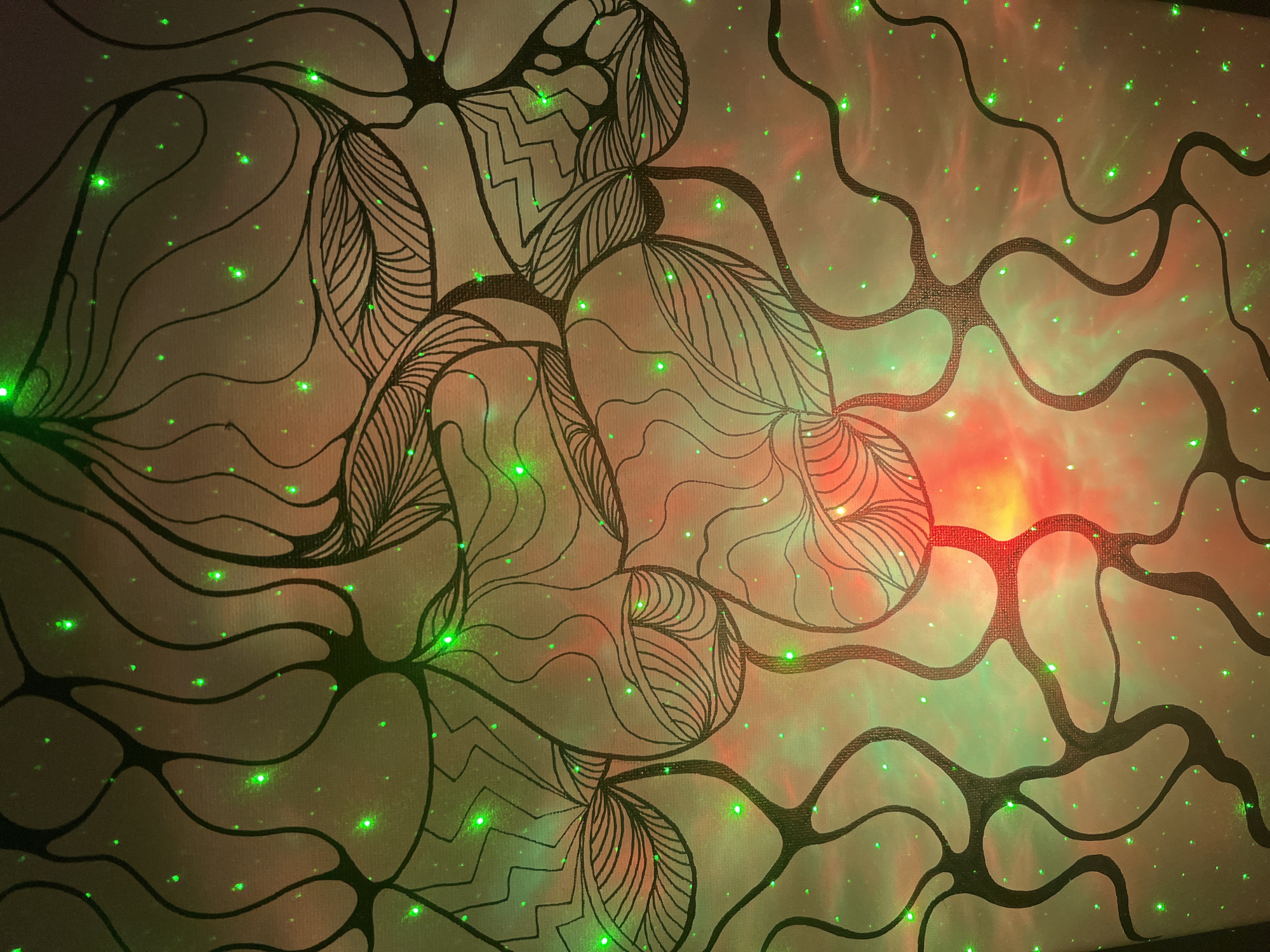 The image displays an abstract indoor scene featuring a light-colored surface, possibly a wall or canvas, covered with a dark, intricate pattern. The pattern consists of thick, flowing, organic lines that intertwine and branch, resembling tangled vines, neural networks, or a complex cellular structure. Within these broader outlines, specific areas are filled with finer line work, including parallel stripes, zigzags, and wavy textures, suggesting stylized leaves or decorative elements.
The surface is illuminated by dynamic and vibrant lights. Numerous small, bright green dots, akin to laser points or scattered stars, are visible across the entire surface. These green points are accompanied by larger, softer glows of colored light. A strong reddish-orange luminescence is concentrated towards the upper and mid-right of the image, blending gradually with a more widespread, diffuse green light that bathes other areas. This interplay of the structured pattern and the ethereal, shifting green and red lights creates a mesmerizing, almost psychedelic, or cosmic atmosphere. No human activity or visible text is present.