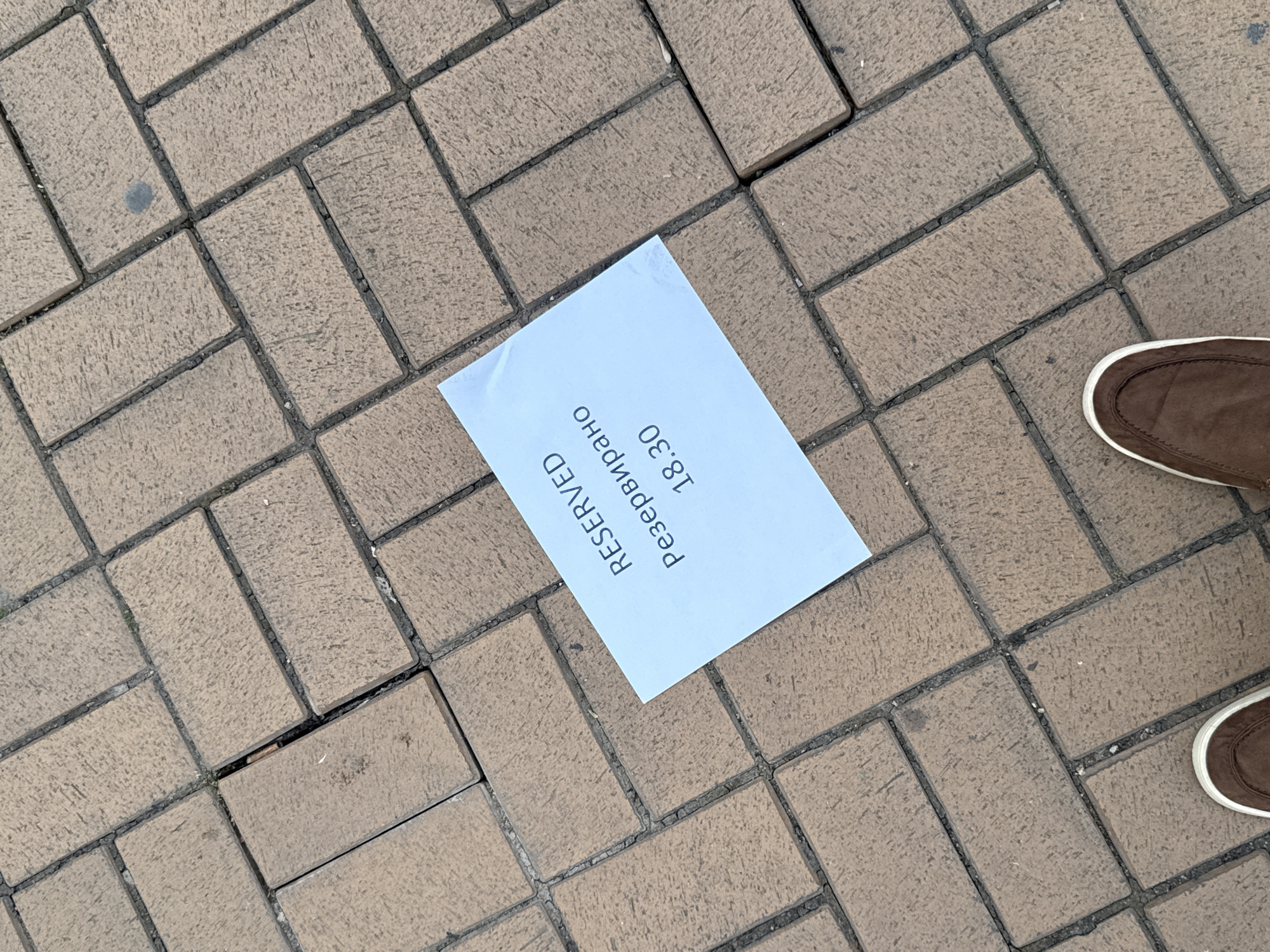 The image shows a paved area constructed of rectangular bricks. In the center of the frame, a white paper with the text "RESERVED" and "резервирано 18.30" printed on it lies askew on the bricks. To the lower right of the frame, the edge of a brown shoe is visible. The pavement is dry. The location is Sofia, Bulgaria.