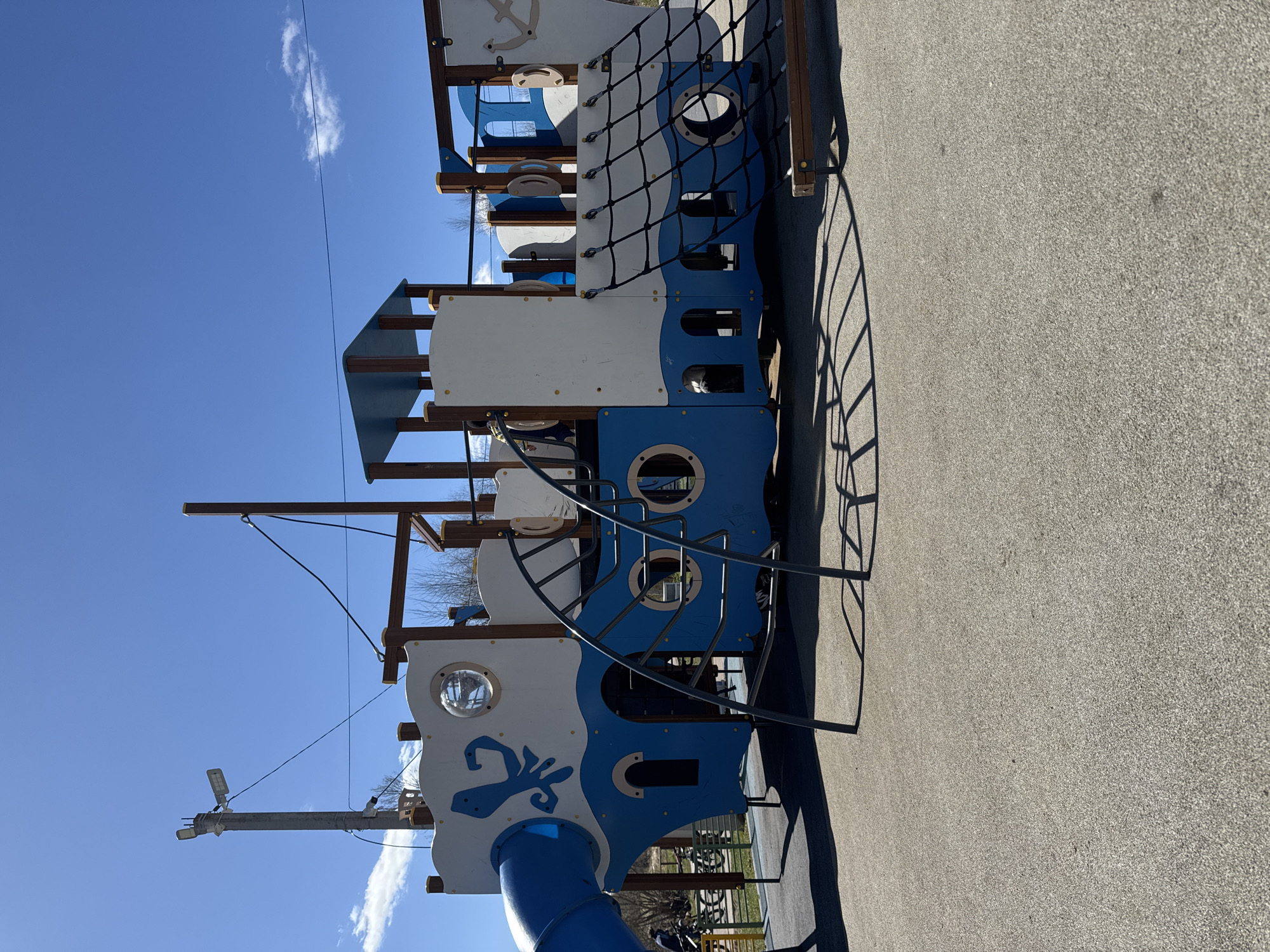 The image shows a large, multi-level playground structure resembling a ship, with blue and white painted panels and nautical-themed decorations. The structure features a slide, climbing nets, portholes, and a ship's wheel. The playground is located on a paved surface. The sky is blue with scattered clouds. The location is potentially Shevlyakovo, Russia, but without further data, that cannot be confirmed from the image itself. There are no people visible in the image.