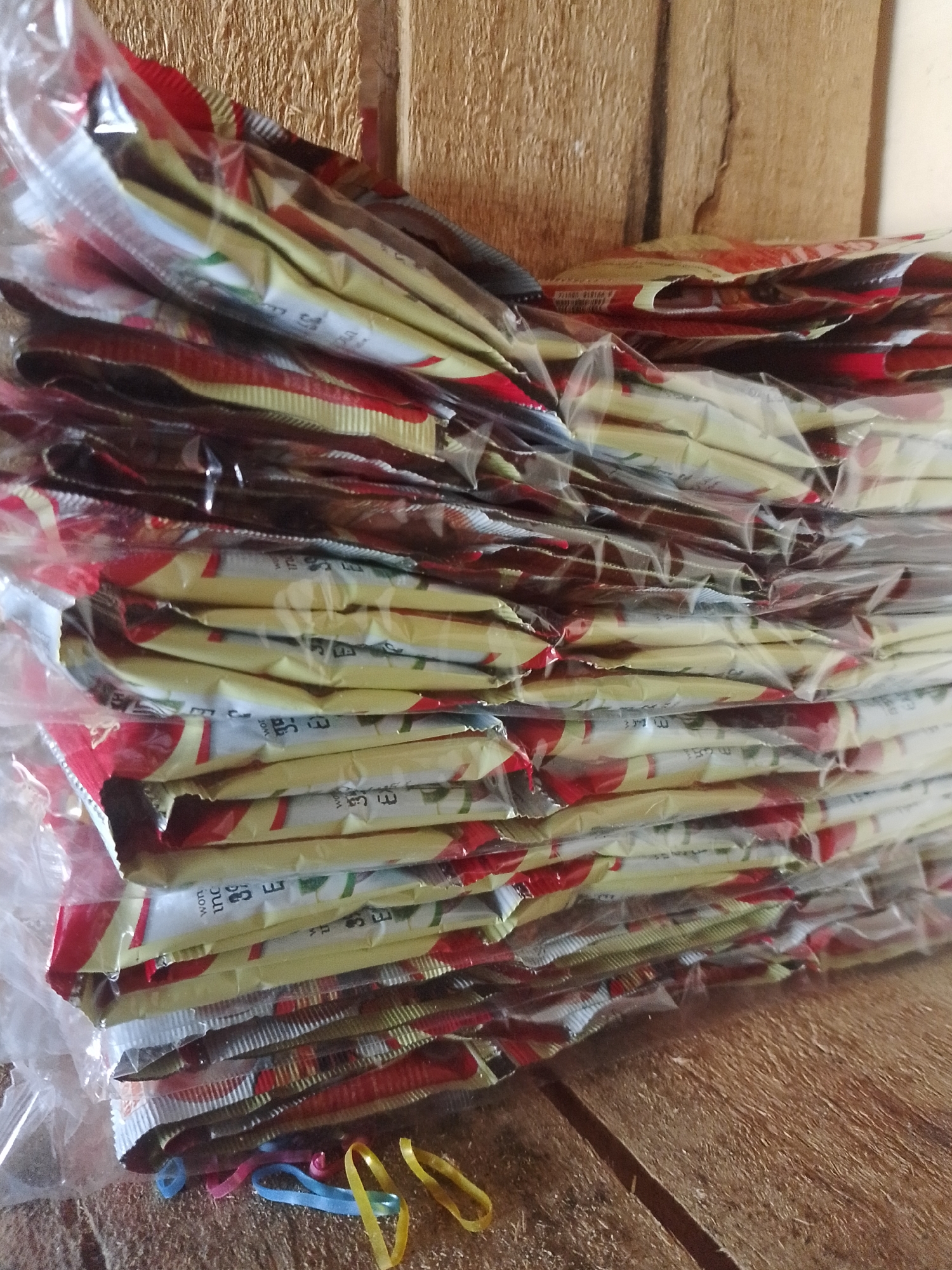 A stack of individually wrapped food packets, likely instant coffee or tea, is piled on a textured wooden surface. The packets are predominantly red and yellow, with some white and green visible. They are enclosed in clear plastic. Three colorful rubber bands, one blue and two yellow, lie on the wooden surface in the foreground. The background consists of weathered, vertically oriented wooden planks. The scene is indoors, possibly in Mukapayung, Indonesia.