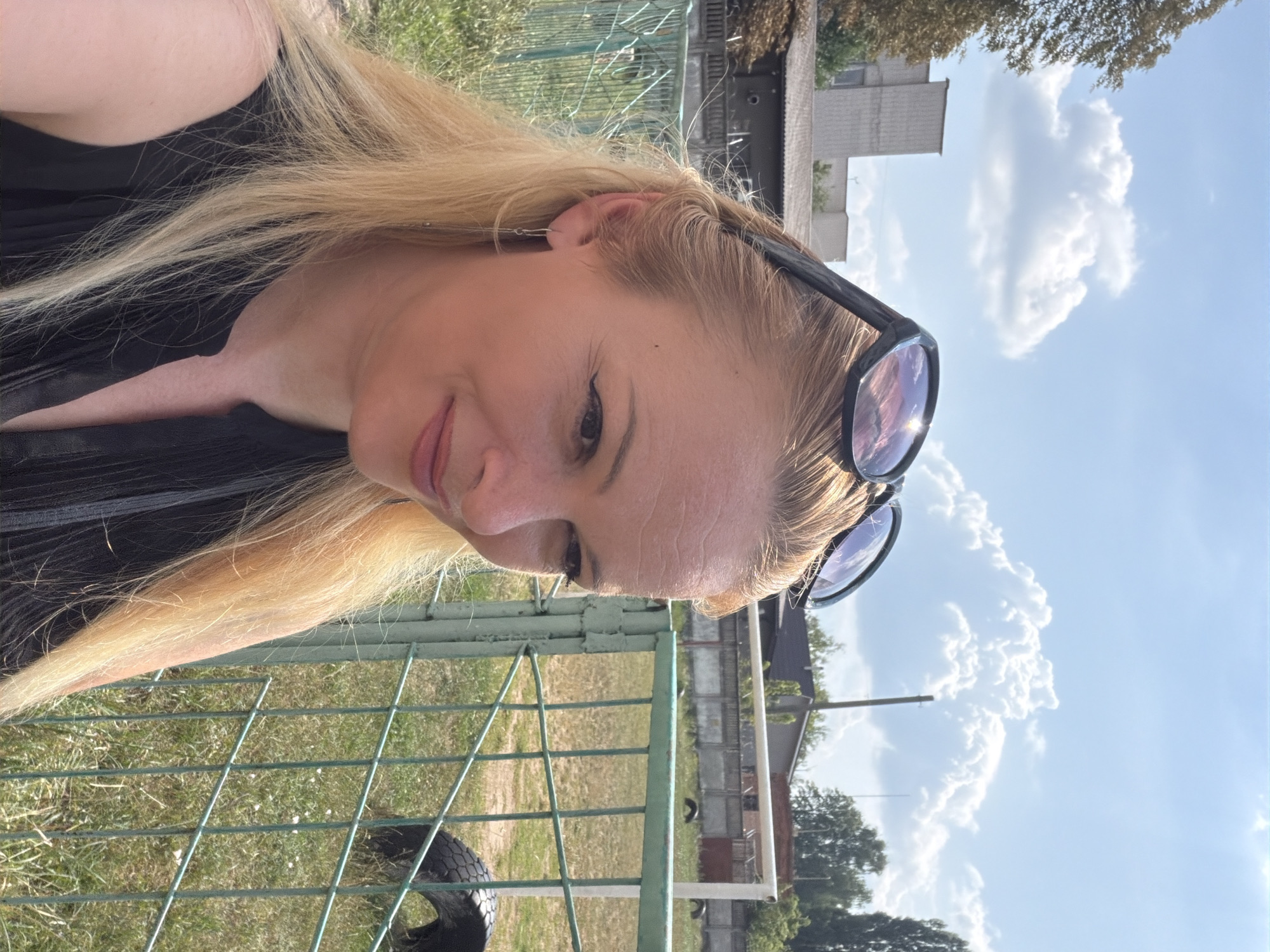 A medium shot shows a fair-skinned woman with blonde hair and sunglasses on top of her head, looking to her right. She is wearing a black top with a pleated V-neck. Behind her, a green metal fence separates her from a grassy field with several old tires scattered on it. In the background, a white soccer goal is visible. The sky is blue with scattered white clouds. Buildings and trees can be seen in the distance. The scene appears to be outdoors in a public area, possibly a park or a sports field.