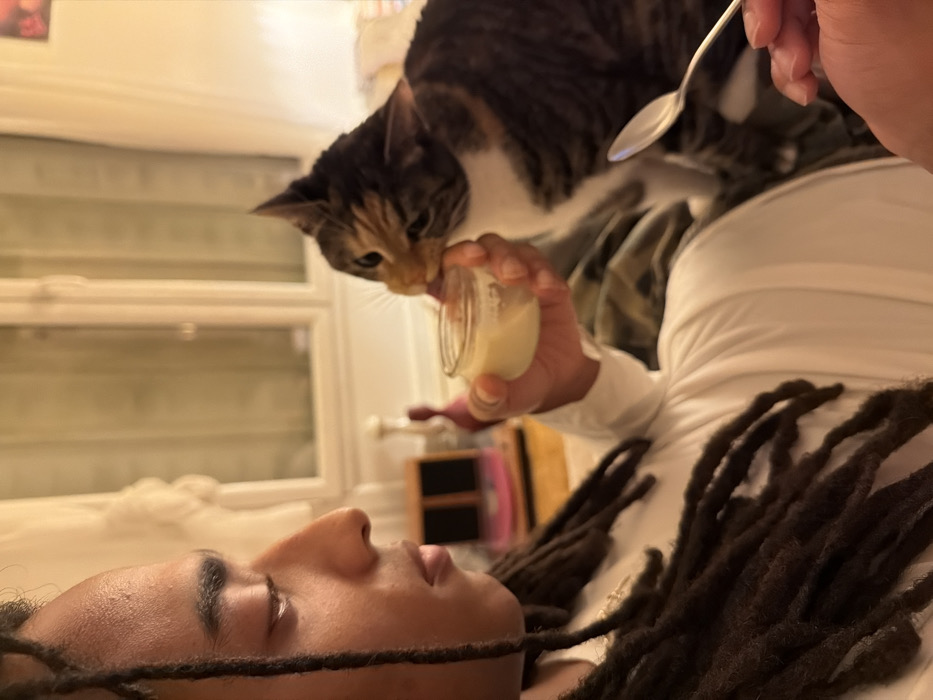 A person with dark skin and long dreadlocks reclines horizontally. Above them, a tabby cat stretches to lick from a small, open jar held in the person's raised hand. The person also holds a spoon. A window with blinds is visible in the background, along with a small wooden cabinet. The person wears a white, long-sleeved shirt. The cat is predominantly brown and white.