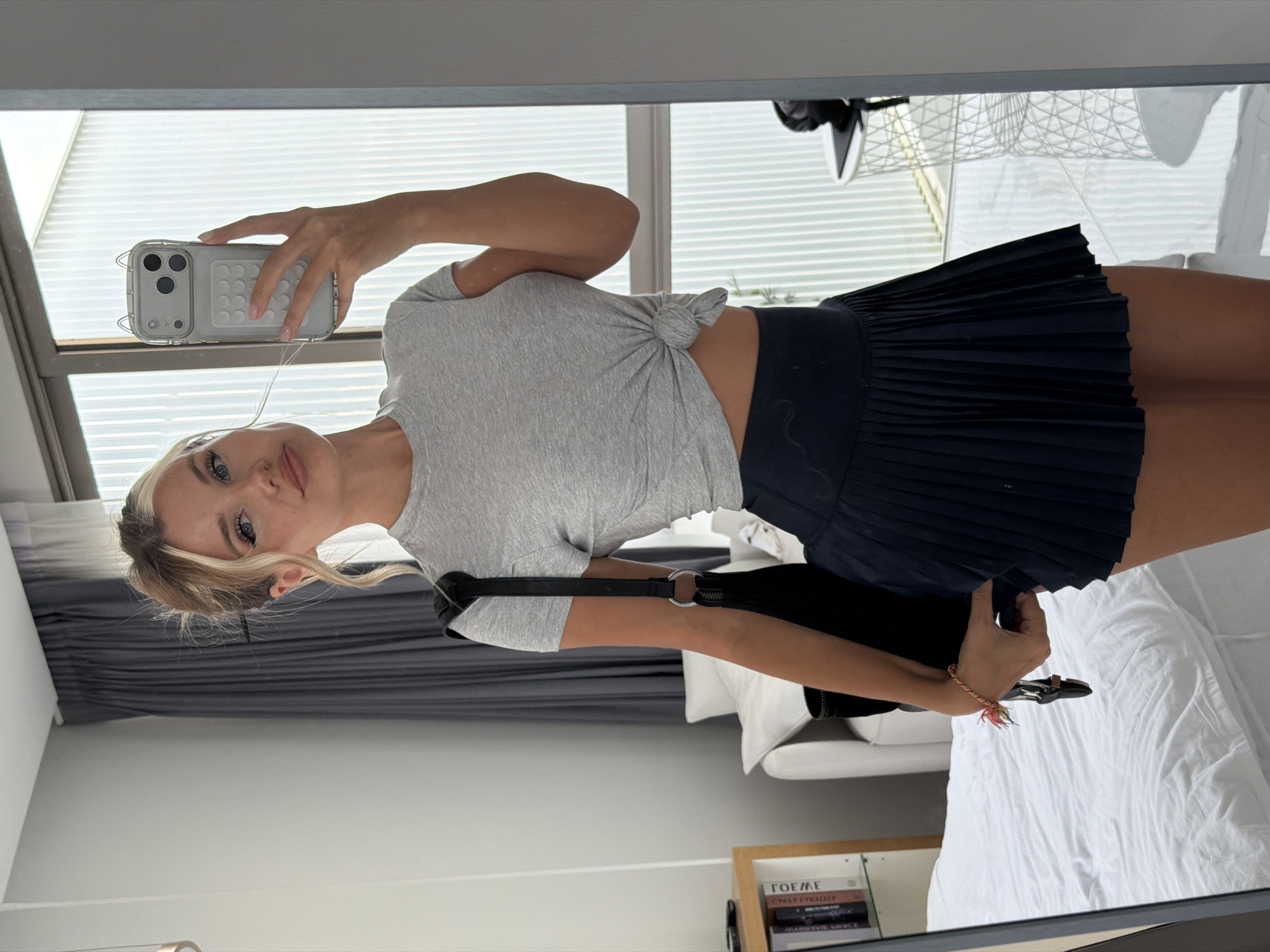 A young Caucasian woman with blonde hair pulled up in a messy bun, wearing a grey, knotted t-shirt and a navy blue pleated skirt, takes a selfie in a bedroom mirror. She is holding a silver iPhone in her right hand and a black handbag over her left shoulder. The room is modern, with a large window to the right reflecting vertical blinds and an outdoor scene. To the left, a white bed and a shelf with books are visible. The location is identified as Thep Krasatti, Thailand.