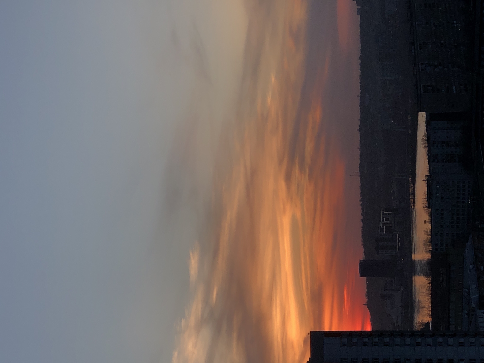 This image, presented in a portrait orientation but depicting a landscape scene, captures a dramatic sunset or sunrise over the city of Krasnoyarsk, Russia.
The sky is the dominant feature, showcasing a striking gradient of colors. The upper left portion of the sky is a calm, muted grey-blue, which transitions towards the right into vibrant, streaky clouds ablaze with intense oranges, reds, and yellows. These fiery hues are characteristic of either a sunrise or sunset, casting a warm glow across the scene.
Below the colorful sky, a dark, silhouetted cityscape stretches across the horizon. Numerous buildings are visible as shadowy masses against the brighter sky, hinting at an urban environment. A body of water, likely a river, horizontally bisects the cityscape, reflecting subtle streaks of light from the sky above. In the foreground, a large, dark building occupies the bottom right, further grounding the urban context of the scene.
The image captures a moment of natural beauty and urban stillness. There are no discernible people or specific activities taking place. The overall mood is one of dramatic serenity, highlighting the contrast between the vibrant sky and the shadowed city below. No text is visible within the image.