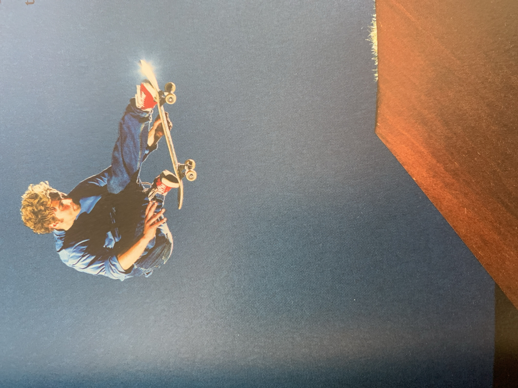 A man in a blue jacket and jeans is airborne, upside down, holding a skateboard over his head, the sunlight glinting off the board. The background is a solid blue color, and a portion of a brown, geometric structure is visible on the right side of the image. The exact location is not specified, and there is no indication it is in Barcelona, Spain.