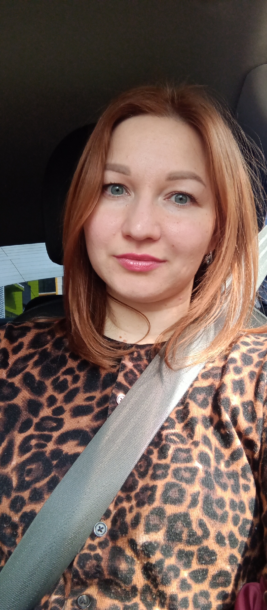 A woman with red hair and light blue eyes is in a car. She is wearing a brown and black leopard print cardigan and a seatbelt. She has light skin with freckles. Part of a building is visible in the background, but its features are indistinct.