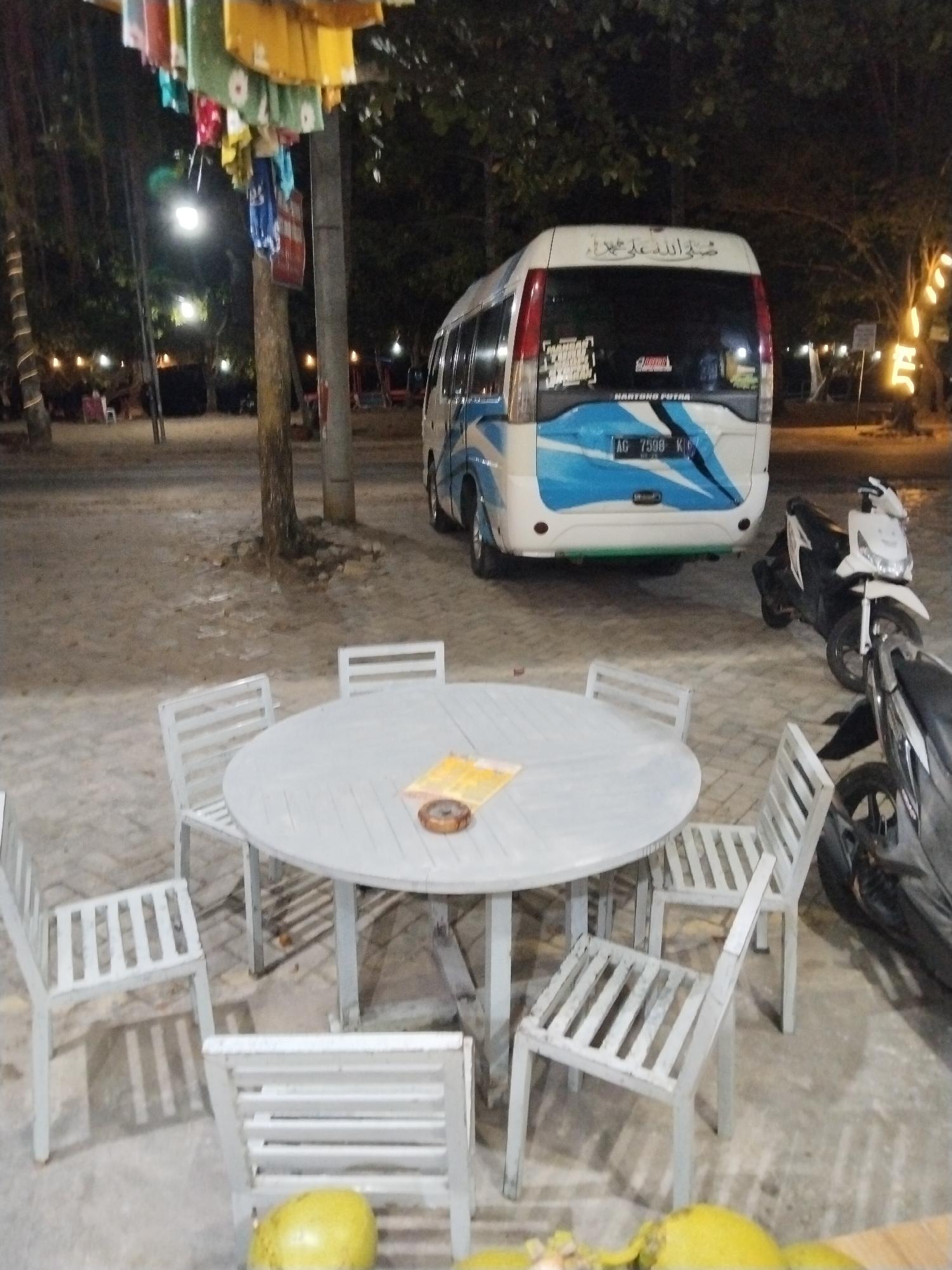 At dusk on a sandy ground in Prigi, Indonesia, a white minivan with blue accents is parked. The minivan has Arabic writing on its rear window and a license plate that reads "AG 7598 K". In front of the minivan, a round white table is set up with chairs. A small pile of menus or pamphlets rests on the table next to an ashtray. Two motorcycles are parked near the table and minivan. In the background, trees and string lights suggest a beachside dining or resting area. There are no people visible in the scene.
