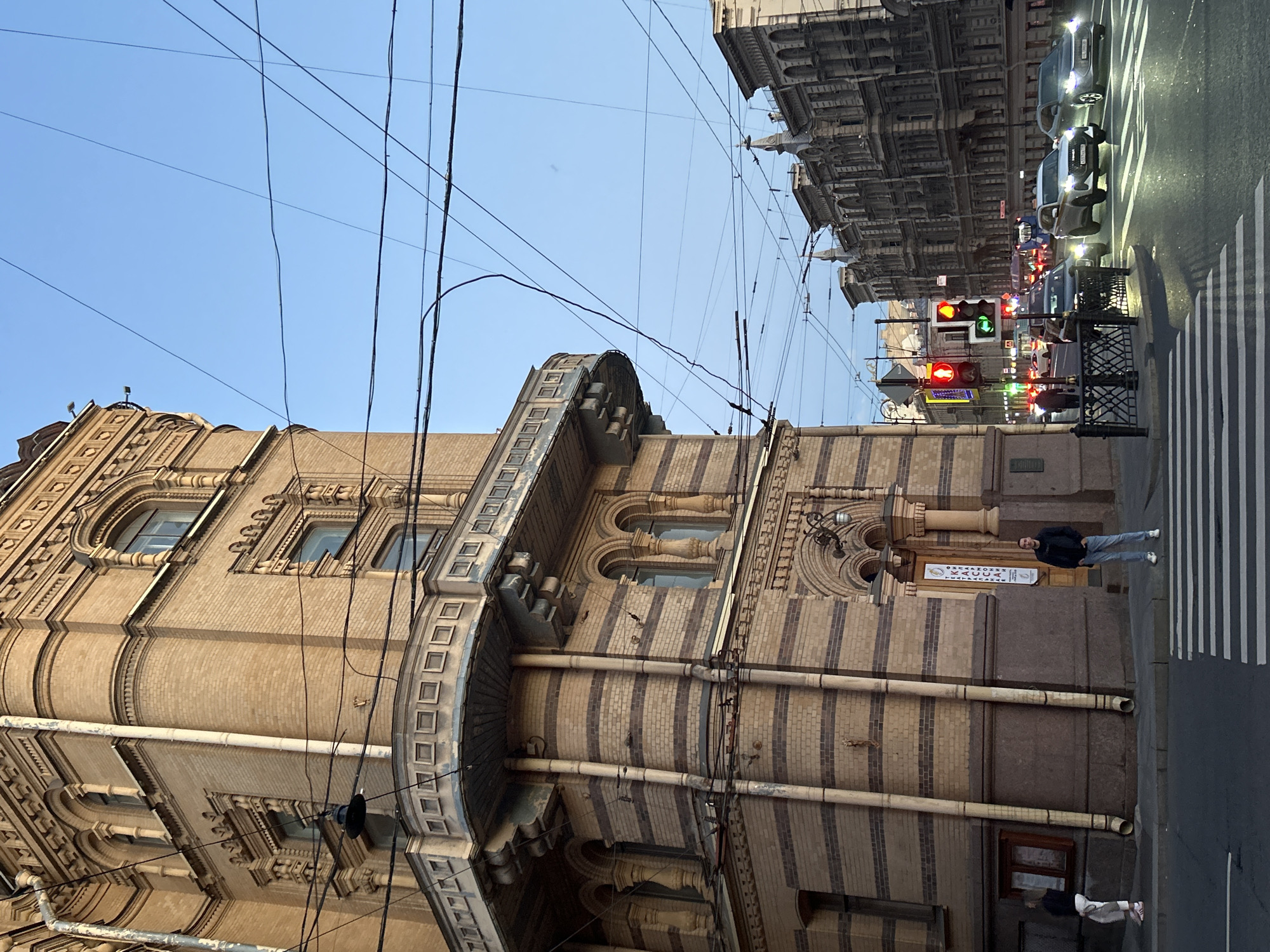A low-angle, full shot captures a street scene in an urban environment, likely at dusk. The left side of the frame is dominated by a large, ornate brick building with a curved balcony. Parallel pipes run vertically along the facade. The building's details include arched windows, decorative brickwork, and what appears to be an entrance with a sign. A man in a dark jacket and light jeans stands on the sidewalk in front of the building, facing the street. Another person, wearing white pants and a dark top, stands further to the left, looking away from the camera. A pedestrian crossing with white stripes is visible in the foreground.
The right side of the image shows a street with multiple lanes of traffic. Several cars are driving on the road, their headlights illuminated. Traffic lights display red, yellow, and green signals. In the background, a row of ornate, classical-style buildings line the street. The sky is clear and blue, with numerous overhead wires crisscrossing the frame. The overall lighting suggests the early evening, with artificial lights beginning to illuminate the street.