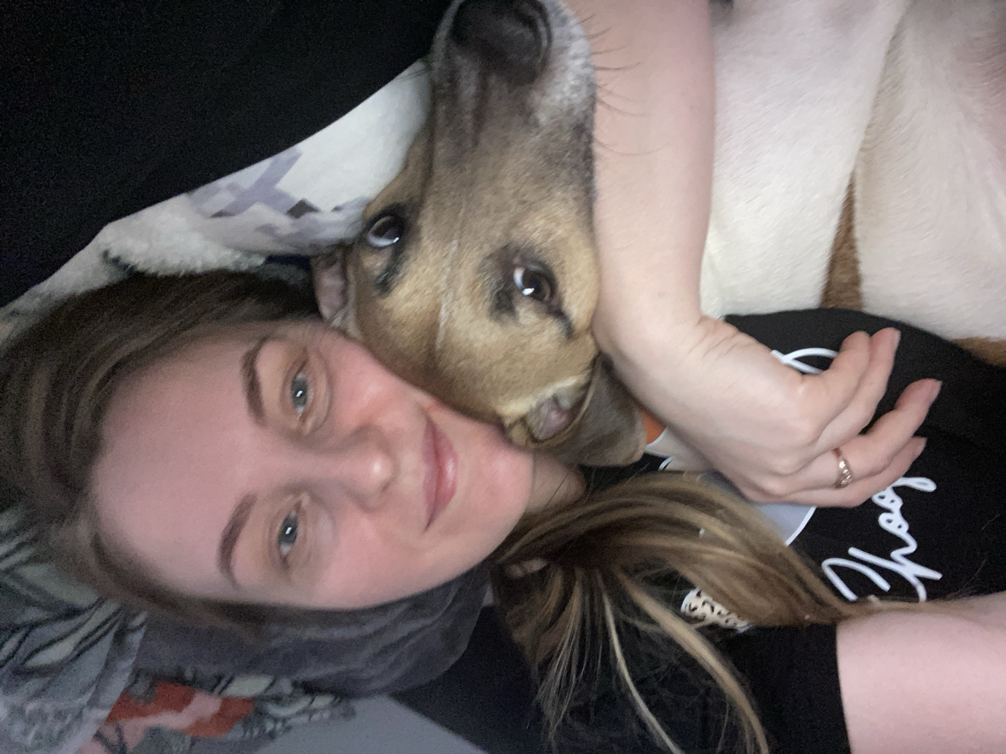 A woman with blue eyes and light brown hair is lying down, looking at the camera. A tan and white dog rests its head on her neck, nuzzling her face. The woman's hand rests on the dog's back. The woman is wearing a black shirt. A ring is visible on her finger. A patterned blanket is behind her. The location is not discernible.