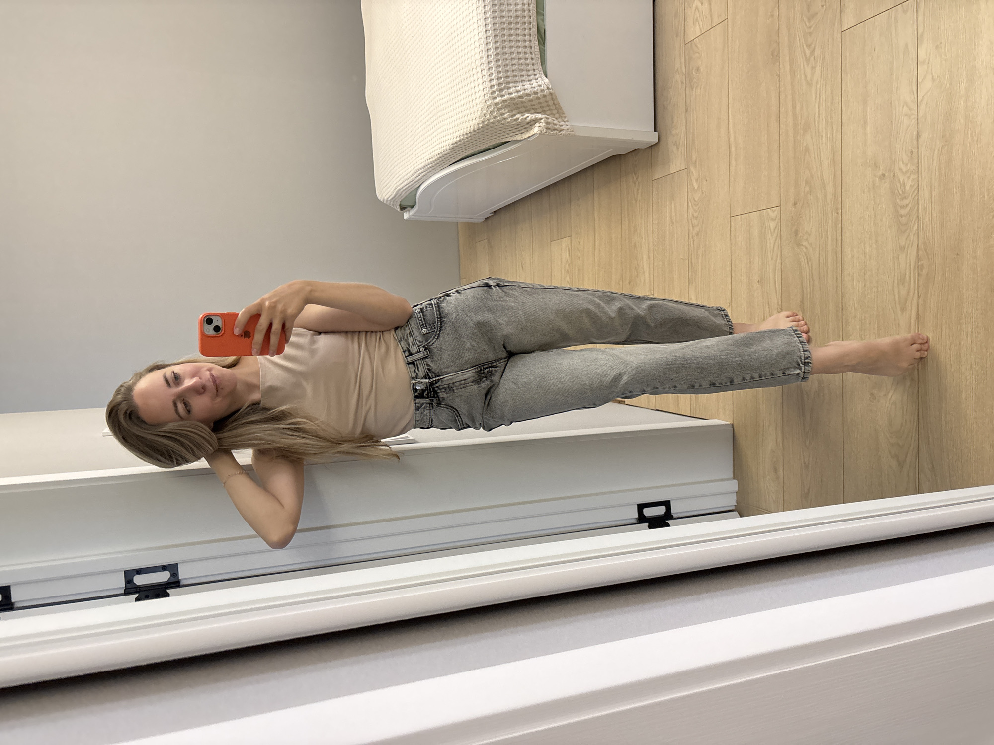 A woman with blonde hair, wearing a beige sleeveless top and grey jeans, stands in a bedroom and takes a mirror selfie. She is holding an orange smartphone in her right hand. Her left hand is behind her head, and her right arm is slightly bent at the elbow. The room has a light-colored wooden floor and a white bed with a textured beige blanket. To the left of the woman is a white doorframe with black hardware. The location is a bedroom in Chaikovsky, Russia.