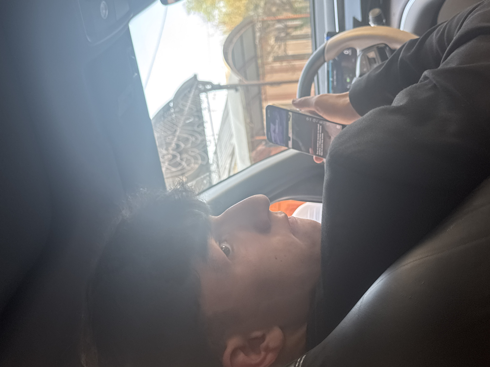 A man is sitting in the driver's seat of a car, holding a mobile phone in his hands. He is looking at the phone screen. The car is moving through a city, evidenced by the buildings and street visible through the windshield and side windows. Based on the architecture and general appearance, it is likely in Tashkent, Uzbekistan.