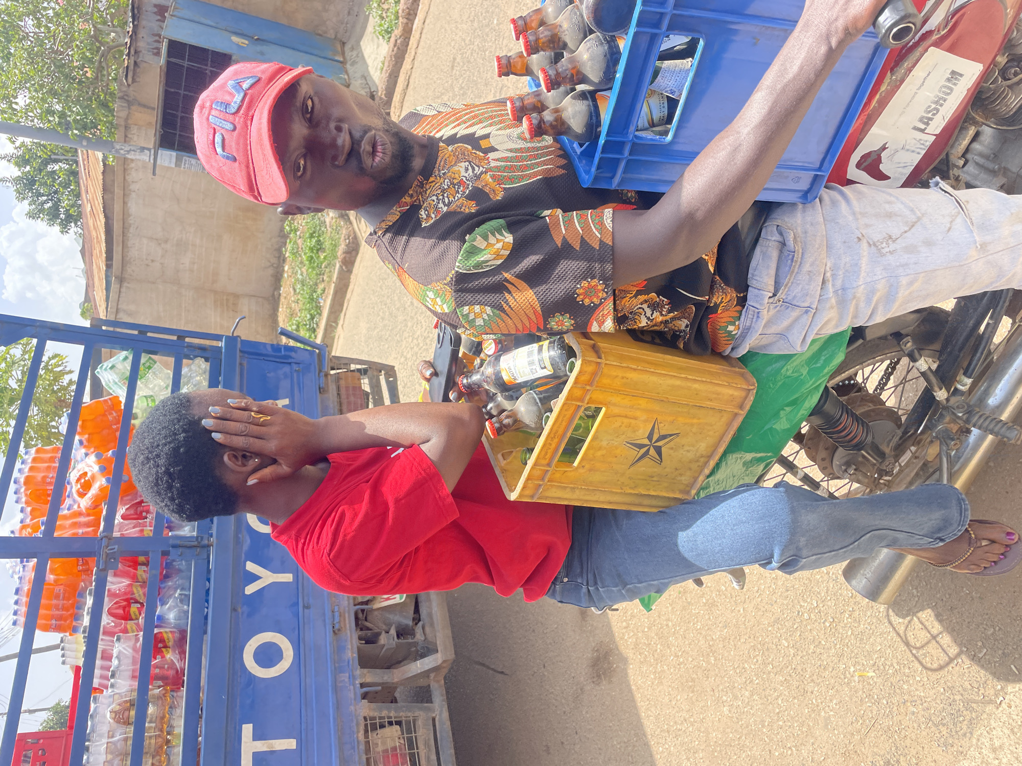 Two individuals are positioned on or near a modified motor tricycle. A man wearing a pink cap and a patterned shirt holds a blue crate filled with bottles while straddling the motorcycle. A woman in a red shirt and blue jeans stands beside the tricycle, holding her hand to her face. A yellow crate containing bottles rests near her. The tricycle's cargo bed is loaded with bottles. The word "TOYO" is visible on the tricycle's blue frame. The setting appears to be a street, with buildings visible in the background. The motorcycle has the word "LASHOM" on its side. The location is Jos, Nigeria.