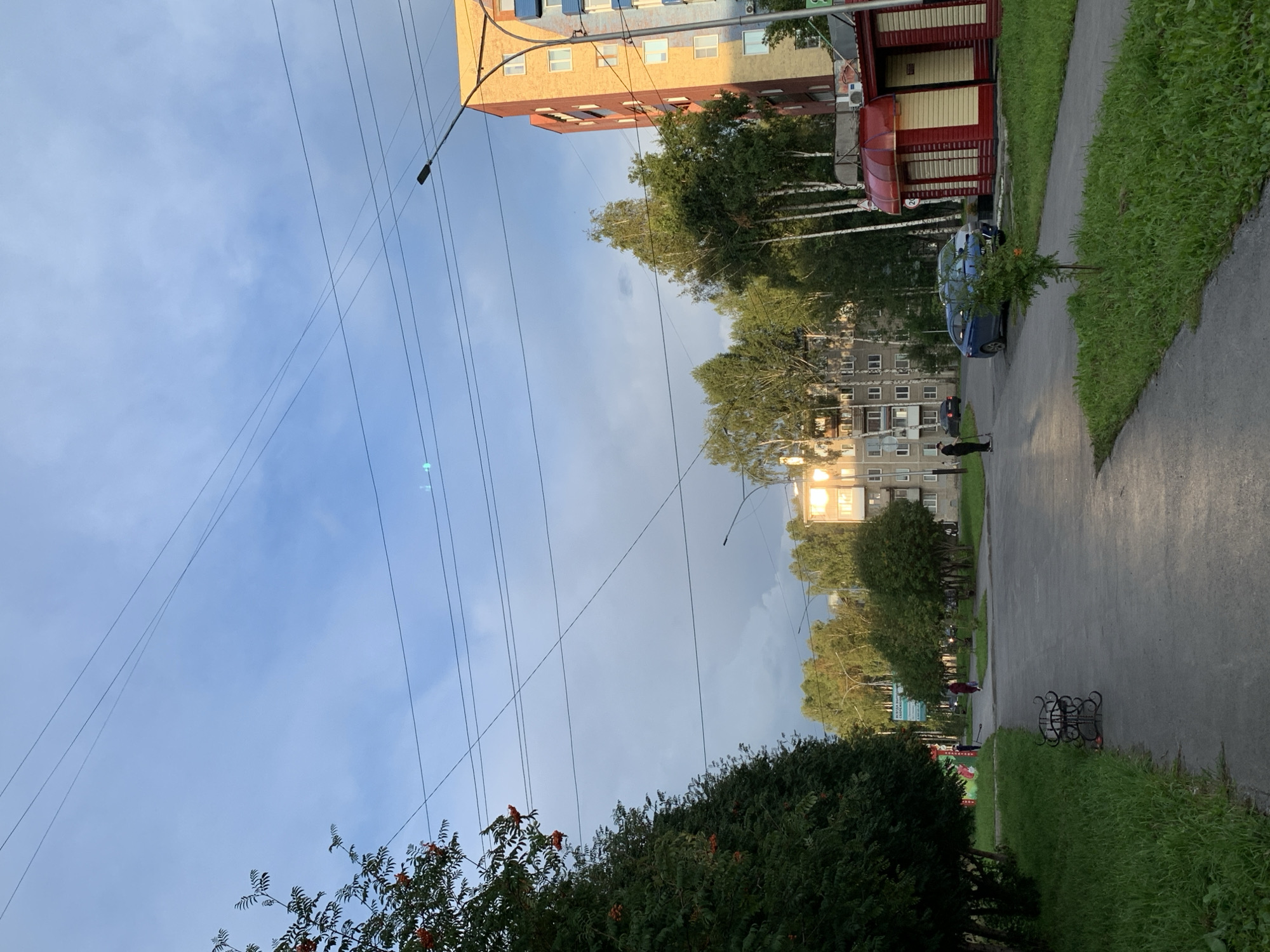 This is a street-level, eye-level shot of a residential area in Prokopyevsk, Russia. The sky is a pale blue with some cloud cover and overhead power lines. On the left, a tree with red berries, likely mountain ash, is visible. In the mid-ground, a paved walkway runs parallel to a road. Several apartment buildings of varying heights and architectural styles are present, with a mix of trees and greenery surrounding them. A few people are visible on the walkway and road. One person is standing near a bicycle rack on the left, and another is walking further down the path. A person on a scooter is visible on the road, along with a blue car and a light-colored car. A small red kiosk with a yellow awning is located on the right side of the image, next to a taller brick building. The lighting suggests it is either early morning or late afternoon, with sunlight casting shadows and highlighting the brick building on the right.