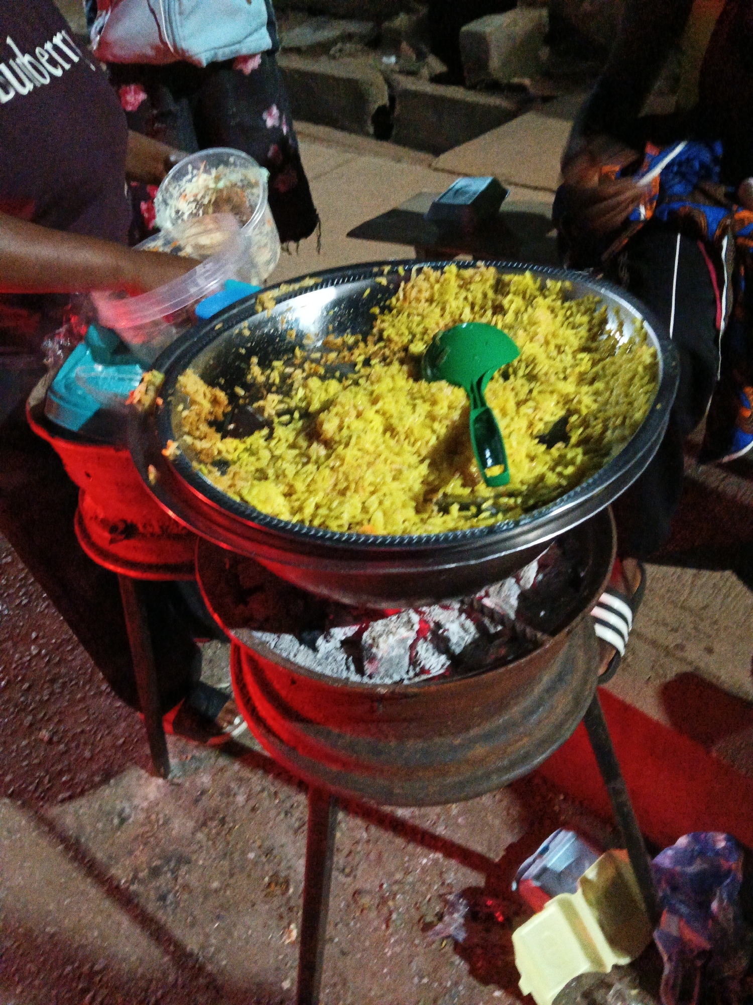 In Bukuru, Nigeria, an outdoor food stall is captured at night. The foreground displays a large, round, metal pot filled with yellow rice, positioned over a charcoal brazier with glowing embers. A bright green plastic ladle rests on top of the rice. To the left, a person's arm and torso are visible, wearing a dark t-shirt with white lettering. Next to the rice pot, a clear plastic container holds a mixture of what appears to be shredded cabbage and other ingredients, possibly a side dish or salad. Behind the stall, indistinct figures and their clothing are visible, suggesting a bustling nighttime environment. Scattered on the ground are various plastic containers and bags. The overall scene depicts a food preparation area set up for nighttime service in an urban setting.