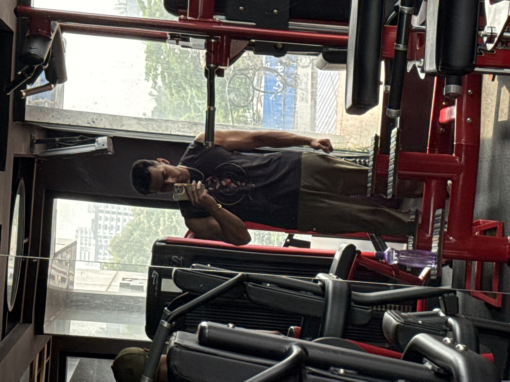 A man is taking a mirror selfie in a gym. He is wearing a black sleeveless t-shirt and olive green pants. He is holding a phone in his right hand. The gym has red and black equipment, including weight machines and benches. Through the large windows, a cityscape with buildings and trees is visible, suggesting an urban setting. The lighting is bright, indicating it is daytime. The man appears to be in good physical shape.