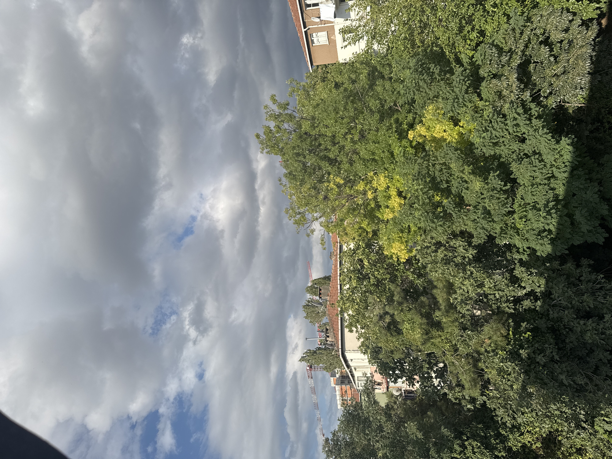 The image captures a view of buildings and trees under a cloudy sky. In the foreground, dense green foliage dominates, with some trees showing patches of yellowing leaves. Behind the trees, several buildings are visible, characterized by reddish-brown tiled roofs and white and brown facades. One building has a satellite dish attached to its exterior wall. In the background, two construction cranes are present, indicating ongoing development. The sky is filled with a mix of dark gray and lighter white clouds, with some patches of blue sky visible. The scene suggests an urban environment in Gungoren, Turkey, on a day with mixed weather conditions.