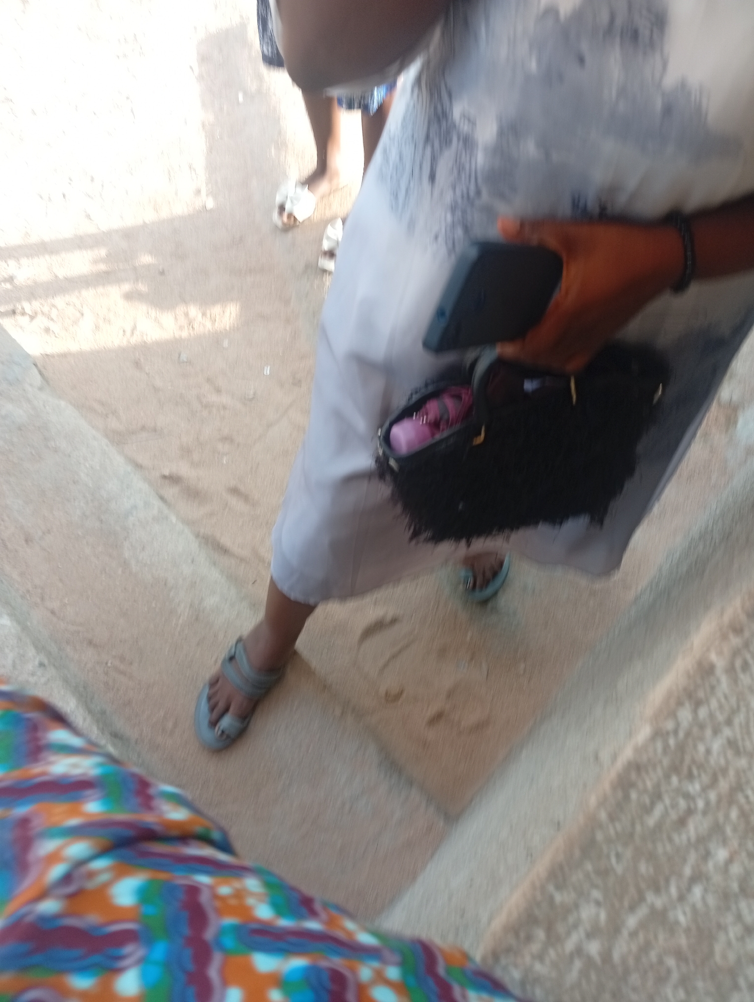 The image depicts a low-angle view of two individuals' legs and feet on a sandy or gravelly surface, possibly a pathway or a dry outdoor area, in Jalingo, Nigeria. The person in the foreground is wearing a brightly colored, patterned garment that obscures much of their body. Their right foot, clad in a grey sandal, is visible. To the left, another person's legs are visible, one clad in a light-colored, possibly white or grey, patterned garment. This person is wearing white flip-flops. A black, textured clutch bag is held in a hand that is partially visible, resting on the light-colored garment. The hand also has a bracelet. The background shows a sandy ground with some shadows, suggesting an outdoor setting. The overall impression is of a casual outdoor scene in Jalingo.